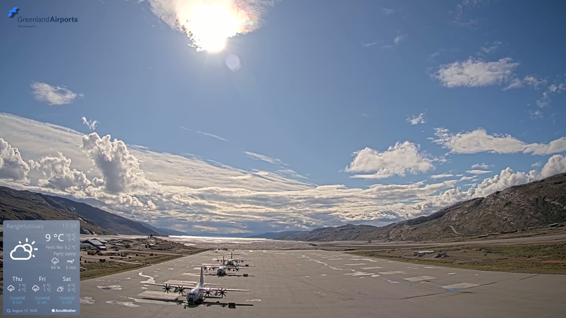 Several airplanes are parked on an airfield under a partly sunny sky with scattered cumulus clouds, and mountains are visible in the background.