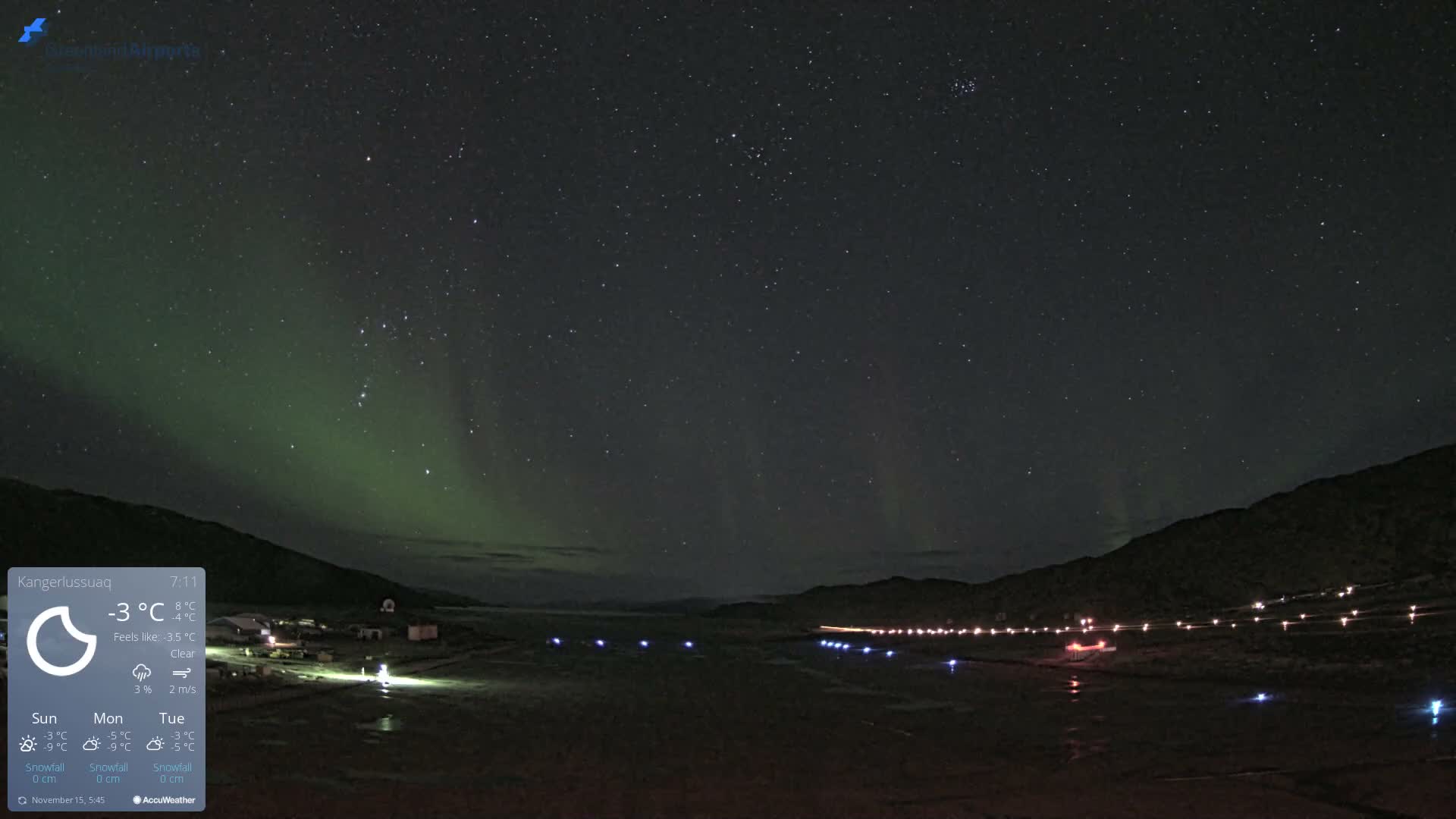 A clear, cold night reveals a vibrant green aurora borealis and a star-filled sky over an illuminated airport runway flanked by dark mountains.