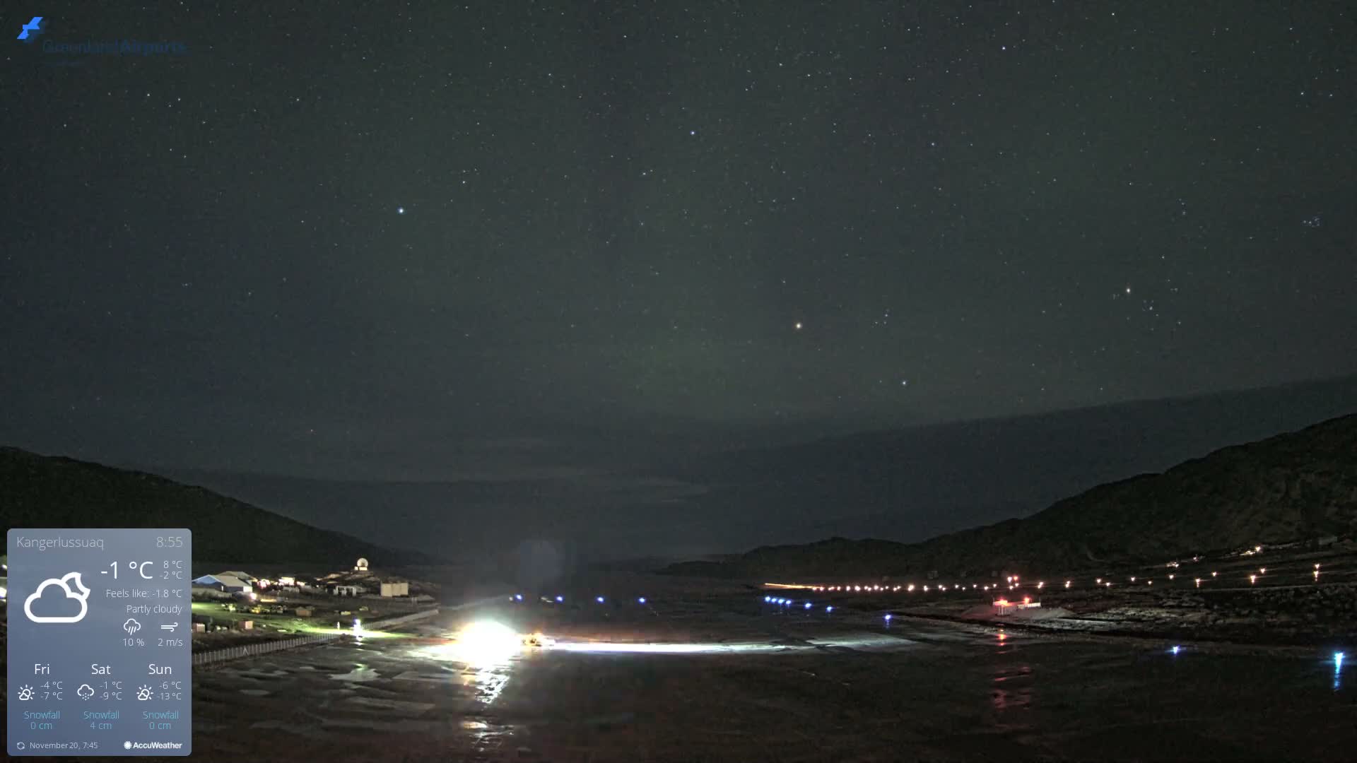 Under a clear, star-filled night sky, a small, brightly lit settlement and a distant runway with blue and white lights illuminate a dark, icy landscape nestled between mountains.