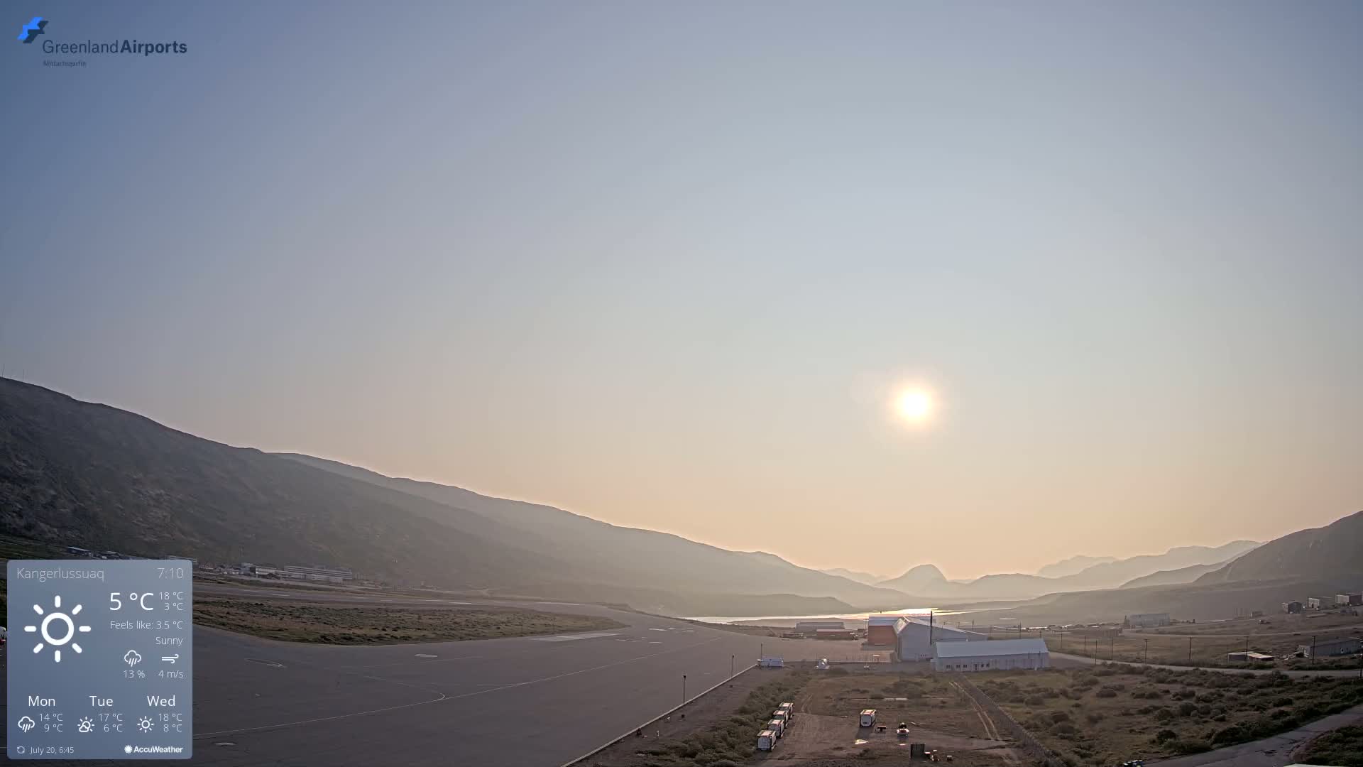A hazy sunrise illuminates a small airport nestled between mountains, with a calm, sunny atmosphere.