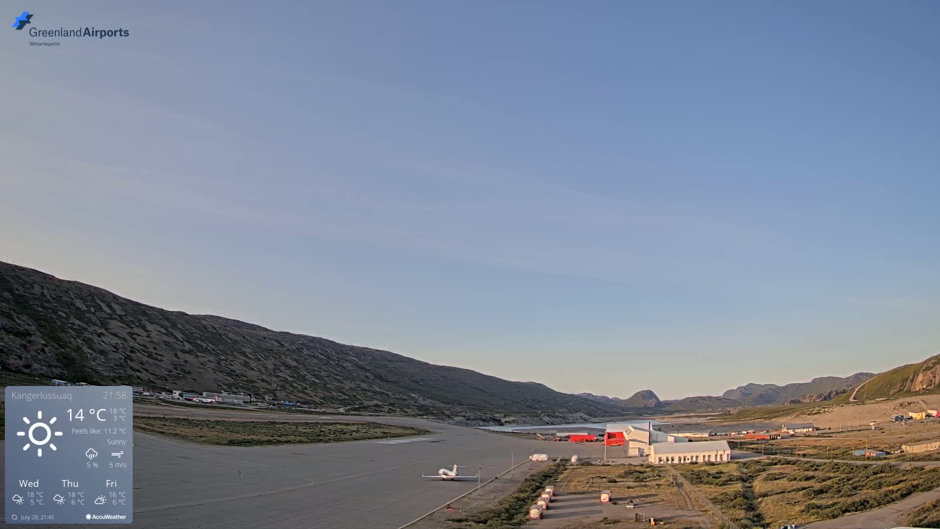 A hazy sunrise illuminates a small airport nestled between mountains, with a calm, sunny atmosphere.