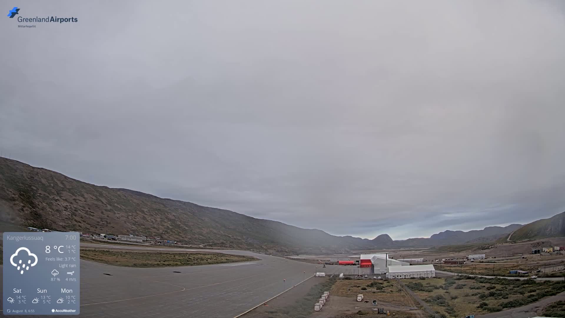 An airport runway and surrounding buildings are visible under a gray, overcast sky with light rain.