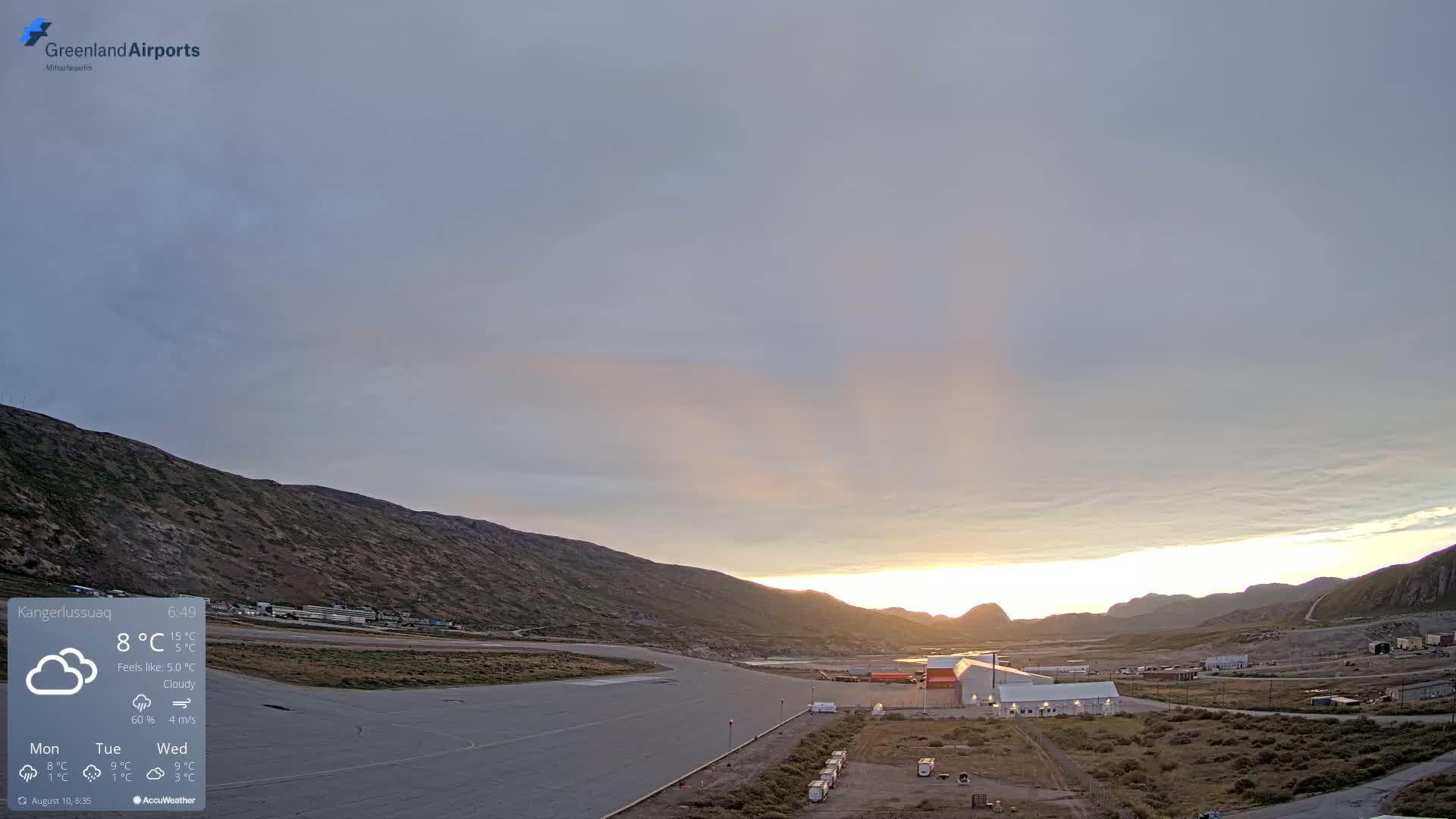 A cloudy sunrise illuminates an airport situated in a valley between rocky hills.