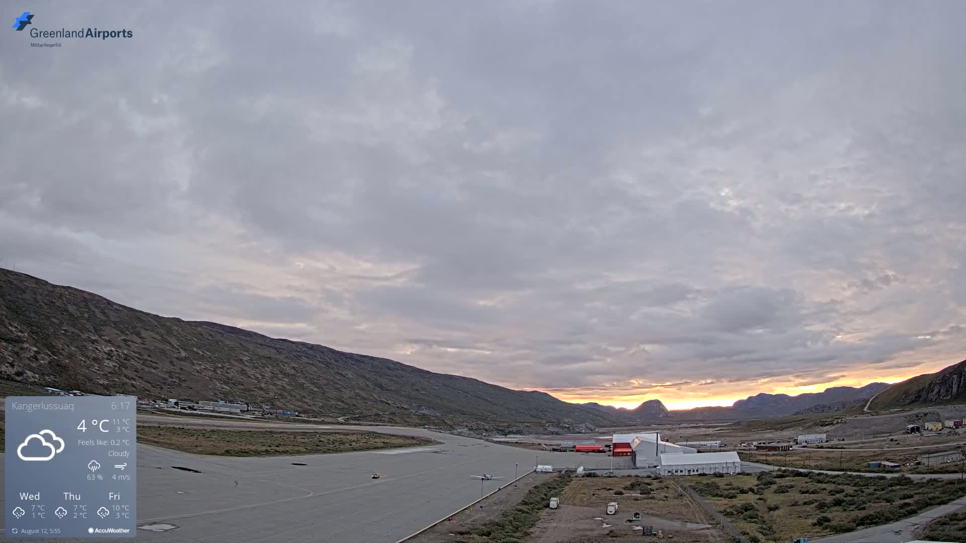 A small airport sits in a valley between low, rocky hills under a mostly cloudy sky with a sunset visible in the distance.