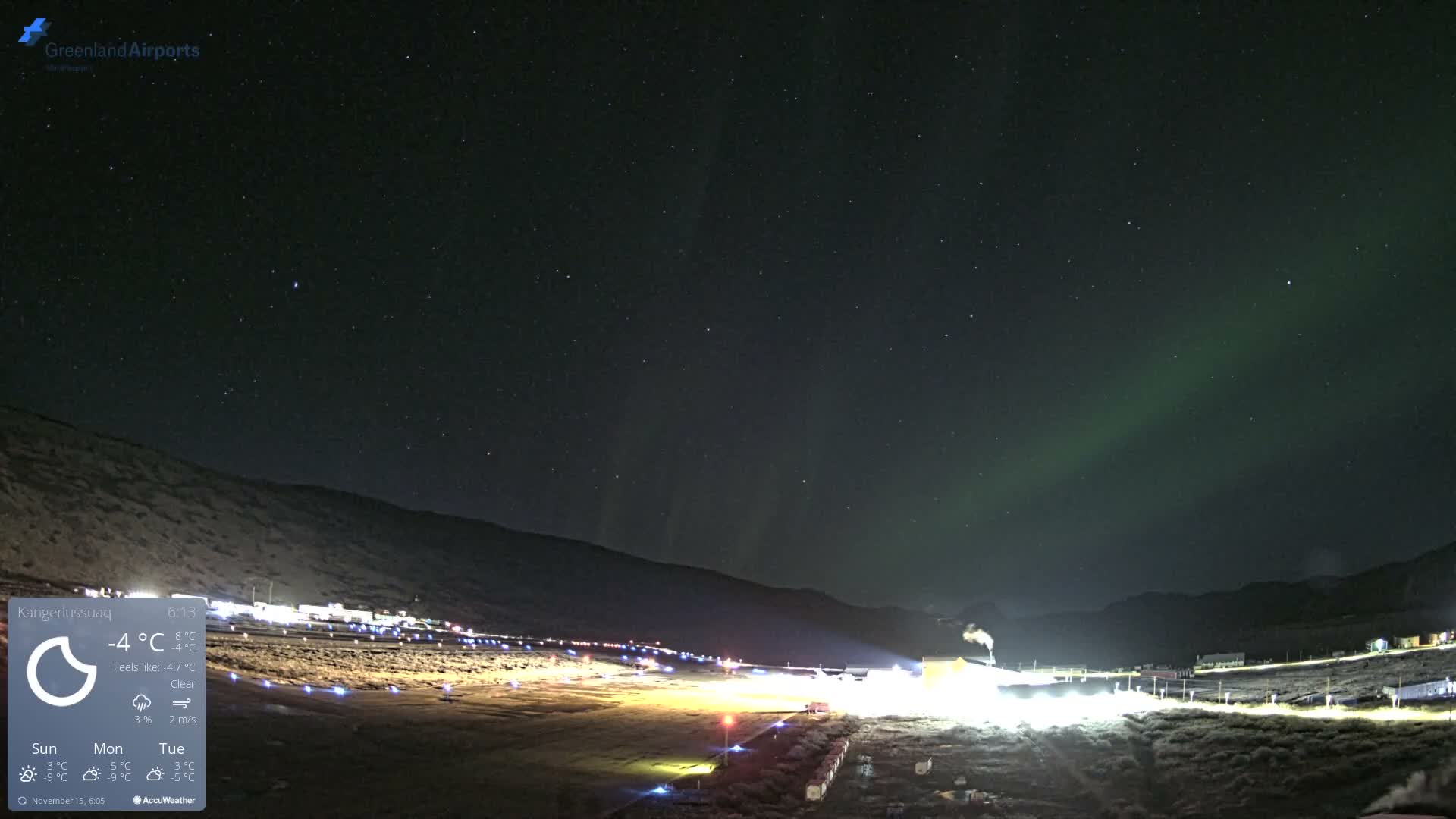 A clear and very cold night sky filled with stars and green aurora borealis stretches above a brightly illuminated ground complex, featuring rows of lights and buildings from which smoke rises, all set against a backdrop of dark hills.