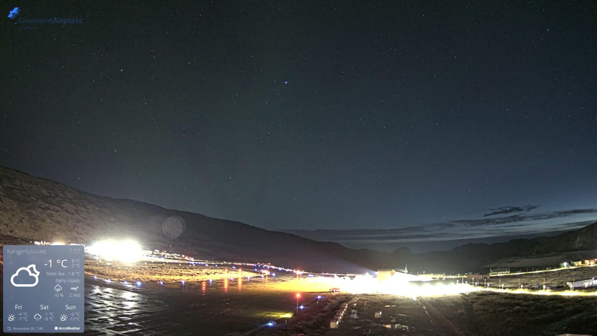 A vast, starlit night sky with some distant clouds hangs over an illuminated airport runway and surrounding structures, reflecting bright lights on a wet or icy surface, all set against a backdrop of dark mountains.