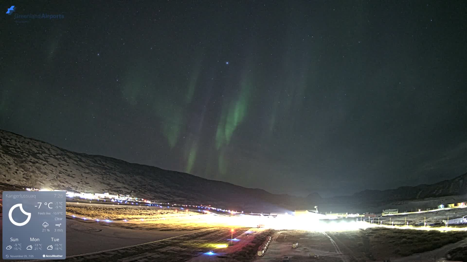The vibrant green aurora borealis streaks across a clear, star-filled night sky above a brightly lit airport runway nestled among snow-covered mountains, indicating cold and clear weather.