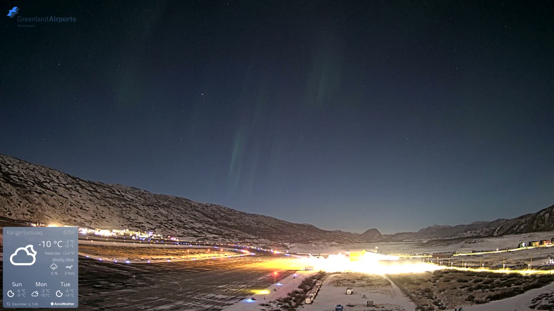 A clear, cold night reveals shimmering green aurora borealis over a snow-covered arctic landscape featuring a brightly lit airport runway and adjacent buildings.