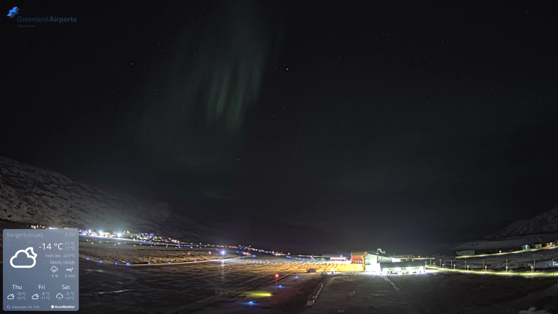 A green aurora borealis shimmers across a starry, dark sky above a snow-covered landscape with an illuminated airport runway and buildings on a clear, cold night.