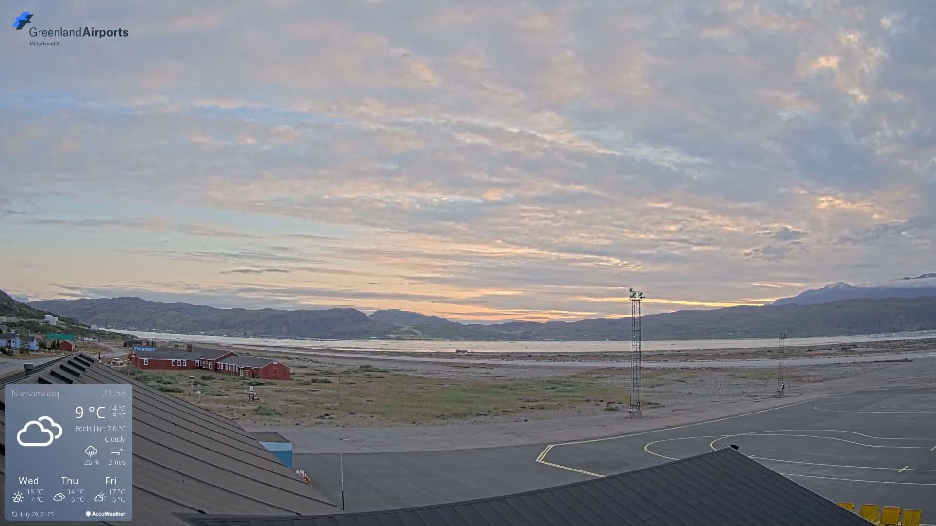 A partly cloudy day reveals a coastal airport with a red building and a body of water surrounded by mountains.