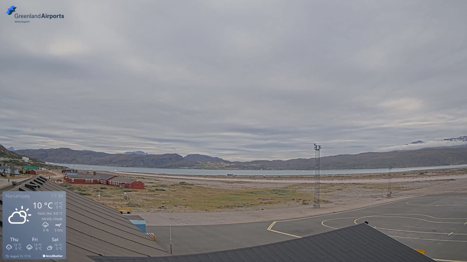 An airport tarmac with a mostly cloudy sky overlooks a body of water and distant mountains.