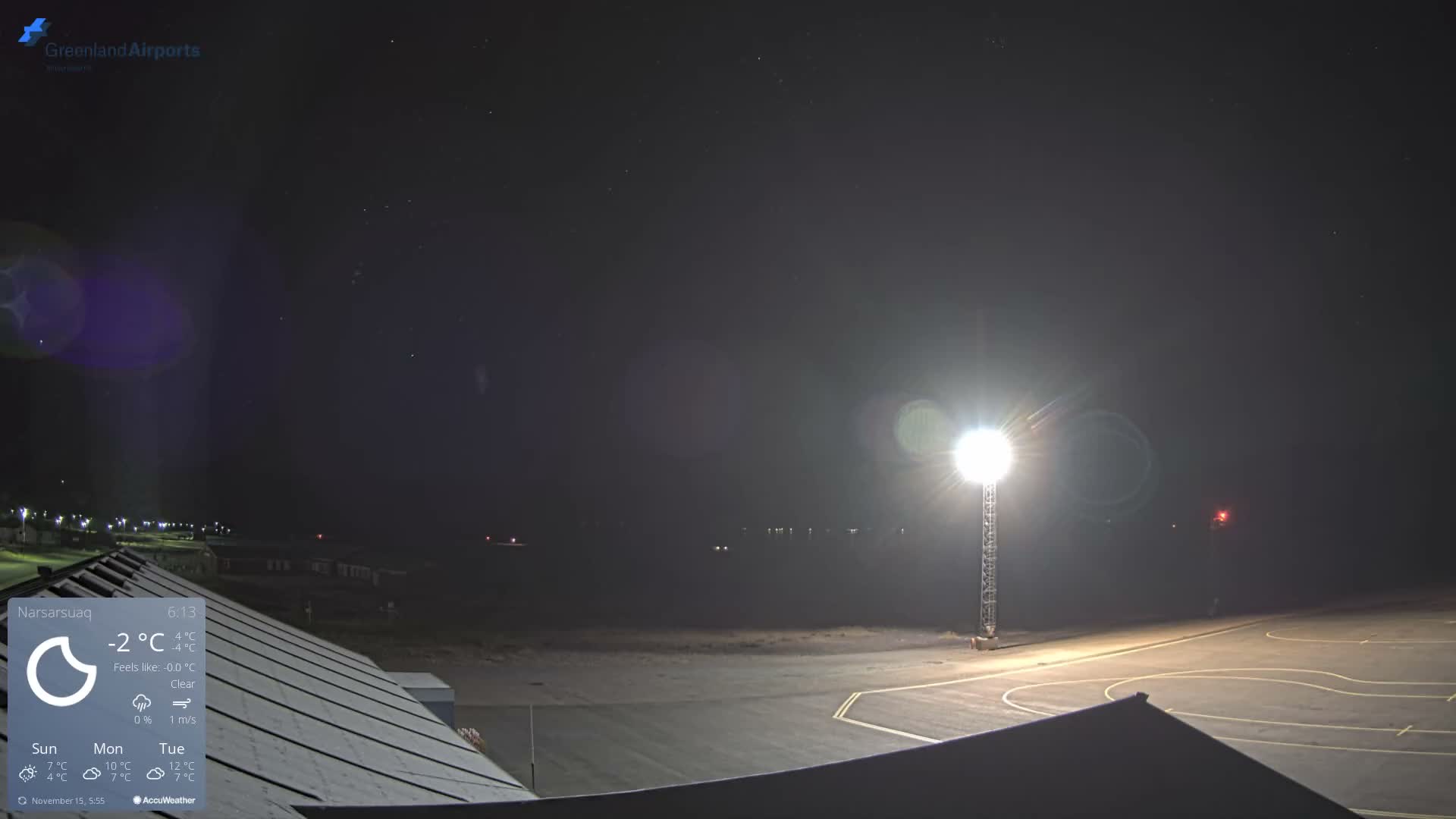 At night, a brightly lit airport tarmac with a building roof in the foreground stretches towards distant lights across a dark expanse under a clear, star-filled sky.