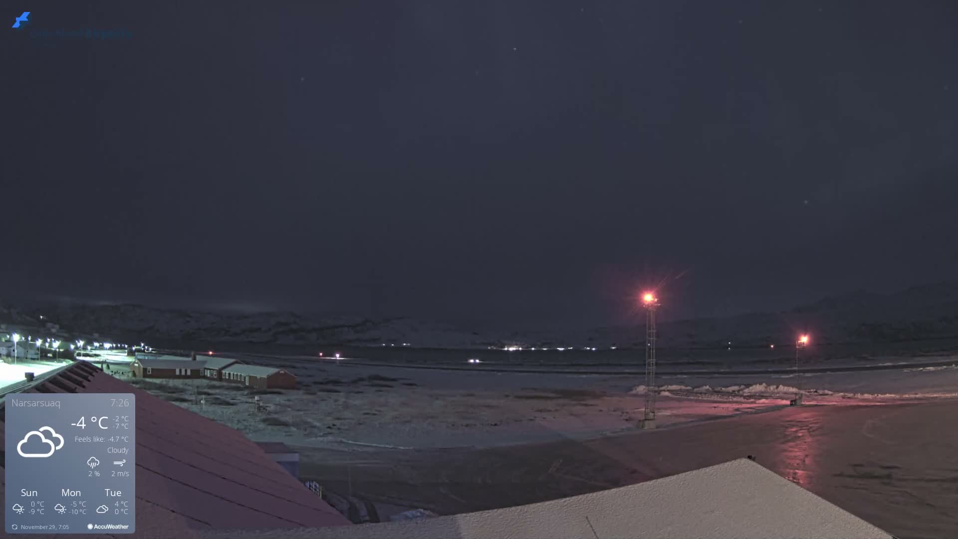 The image depicts a snowy landscape at night, featuring bright red lights illuminating an open area that appears to be a runway, surrounded by buildings and distant dark mountains.