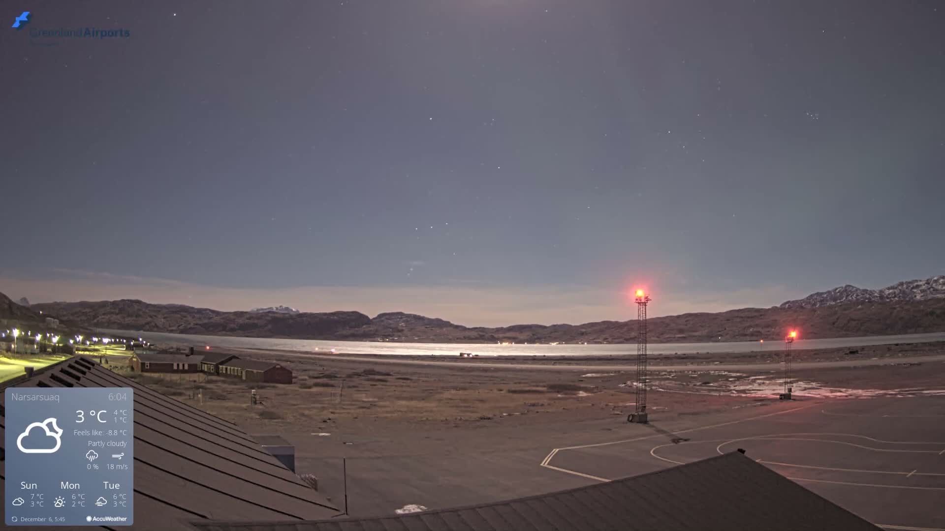A nighttime view reveals a dark airfield with two prominent red-lit towers, a calm body of water, distant village lights, and rugged mountains under a clear, star-dusted sky.