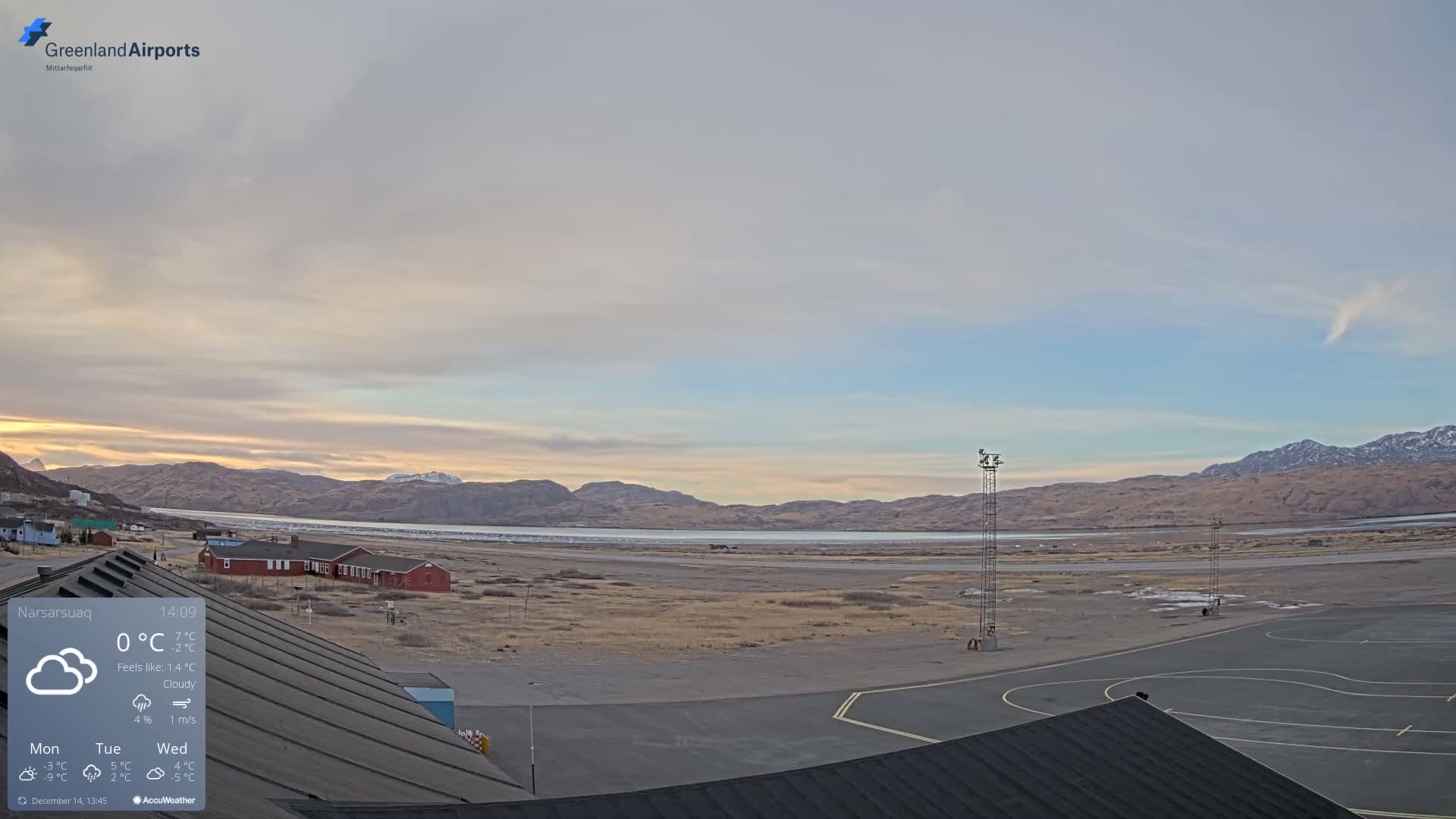 A nighttime view reveals a dark airfield with two prominent red-lit towers, a calm body of water, distant village lights, and rugged mountains under a clear, star-dusted sky.