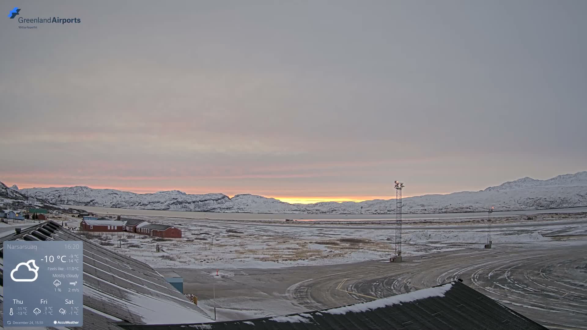 A vast, snow-covered and cold landscape stretches under a dark, partially cloudy, starry night sky, illuminated by red warning lights and distant town lights amidst surrounding mountains.