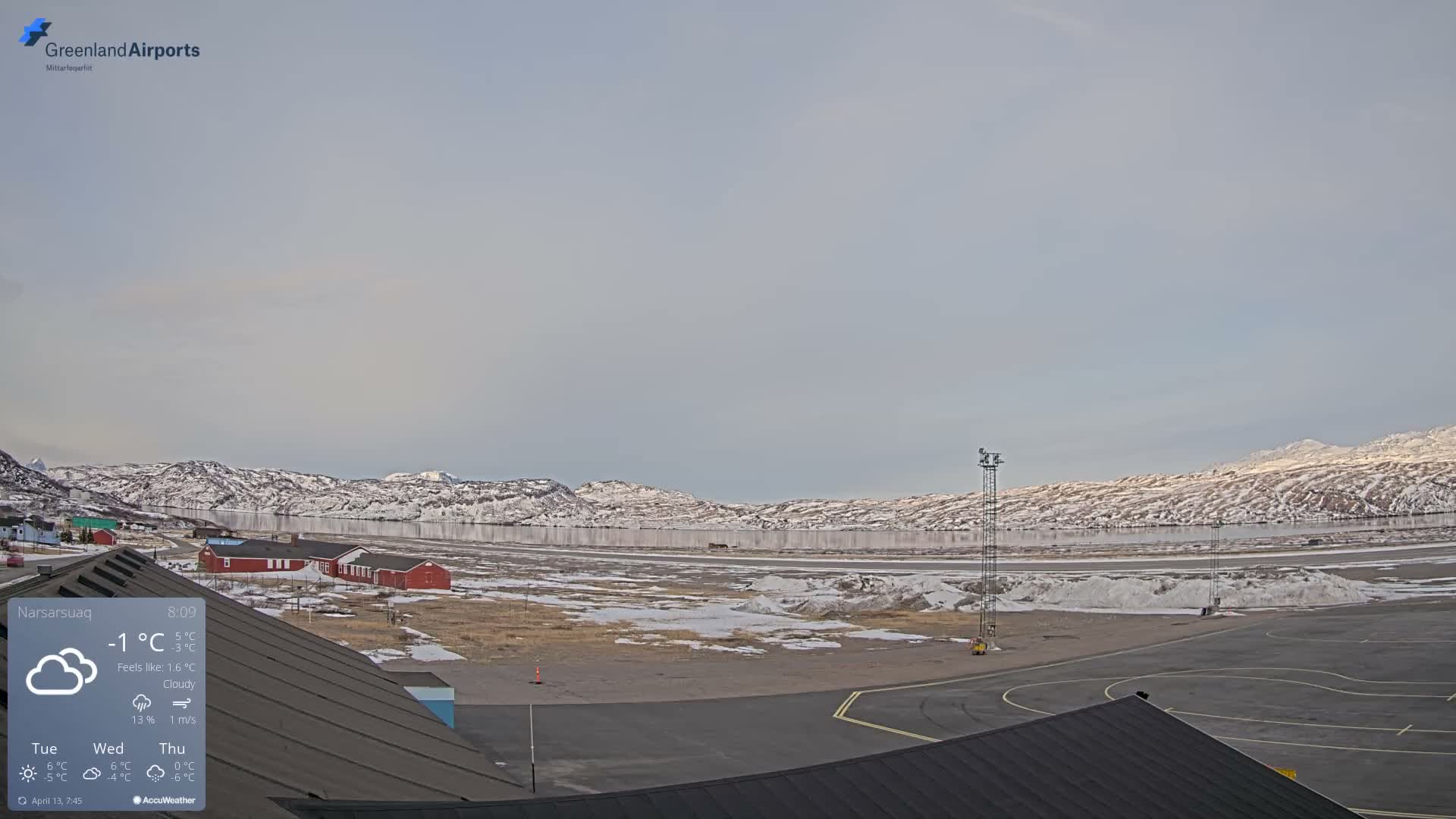 A vast, snow-covered and cold landscape stretches under a dark, partially cloudy, starry night sky, illuminated by red warning lights and distant town lights amidst surrounding mountains.