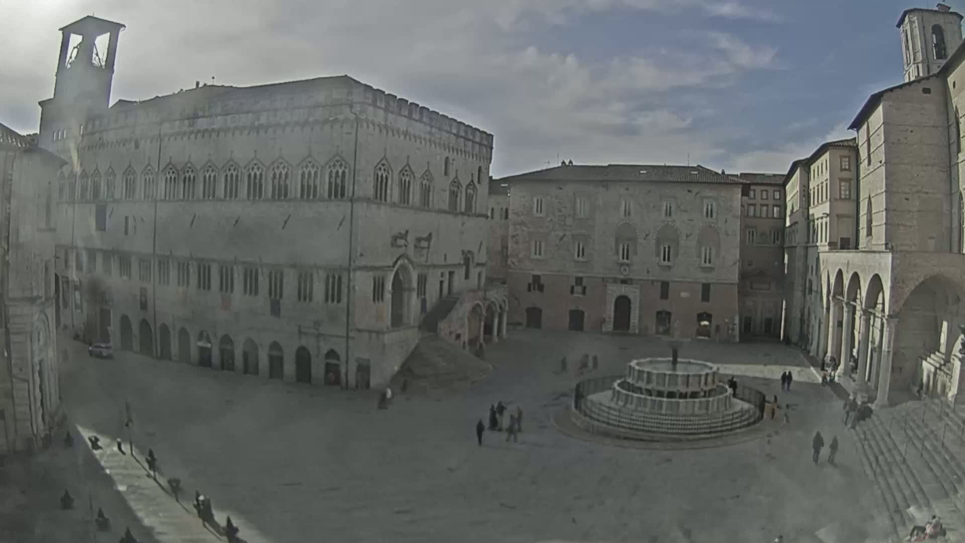A large, mostly empty town square with a fountain at its center is surrounded by several aged stone buildings under a cloudy sky.