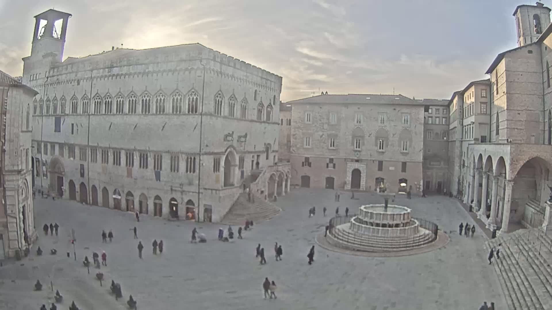 A large town square with several stone buildings, a fountain, and a scattering of people, under a mostly cloudy sky.