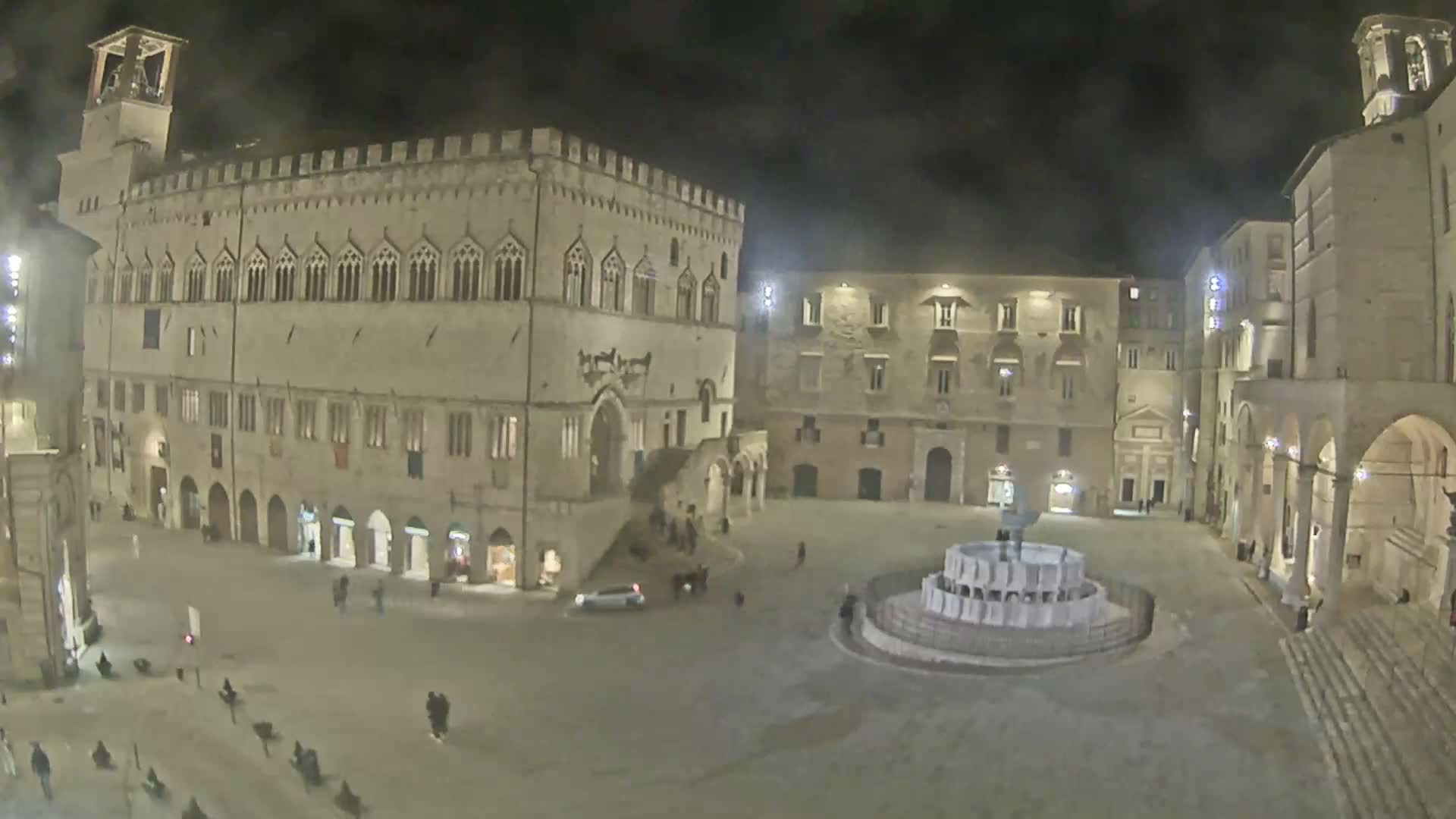 A nighttime view of a large, mostly empty town square surrounded by ancient stone buildings, featuring a fountain in the center.