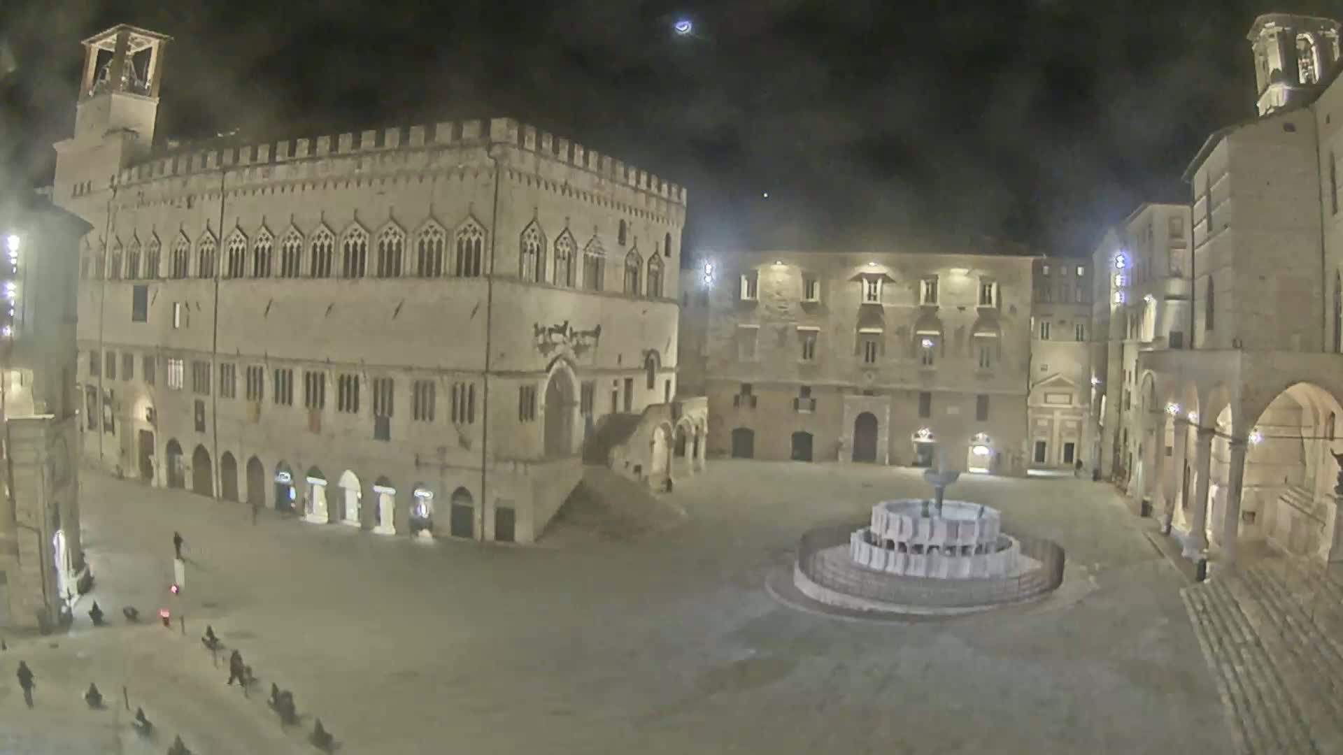 A nighttime view of a town square with a large stone building and fountain, under a dark sky.