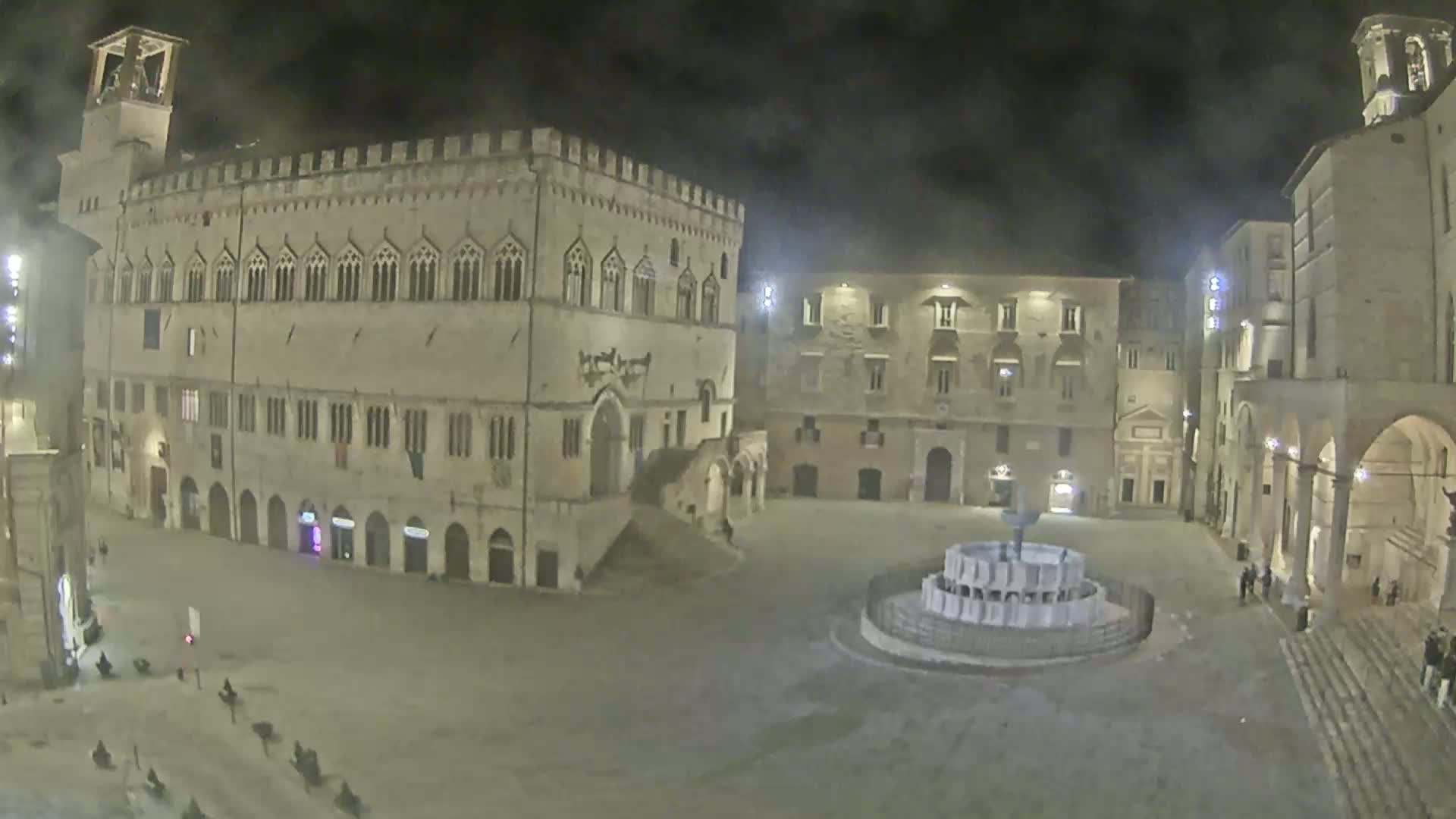 A nighttime view of a large, mostly empty town square with several historical buildings surrounding it, including a large palazzo, and a fountain at the center.
