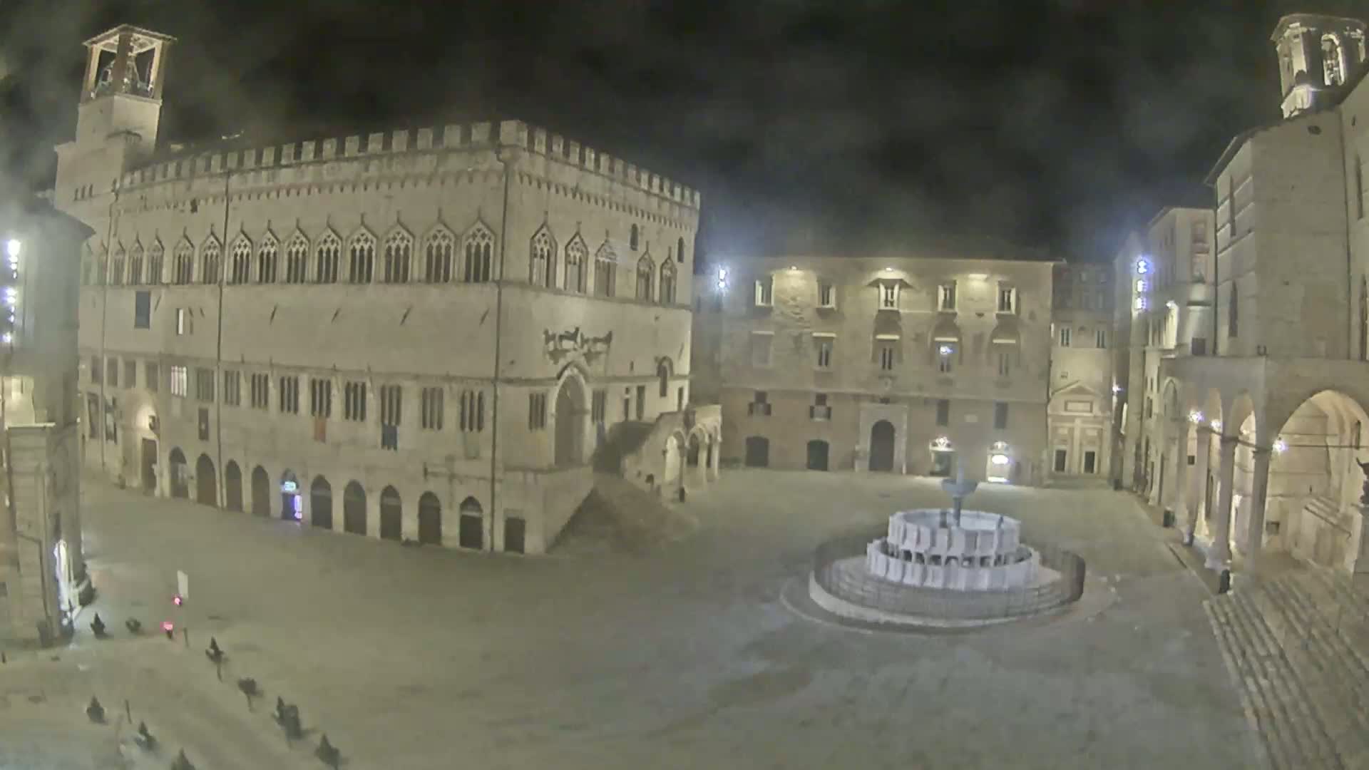 A nighttime view of a large, mostly empty town square with a fountain at its center, surrounded by historic stone buildings.