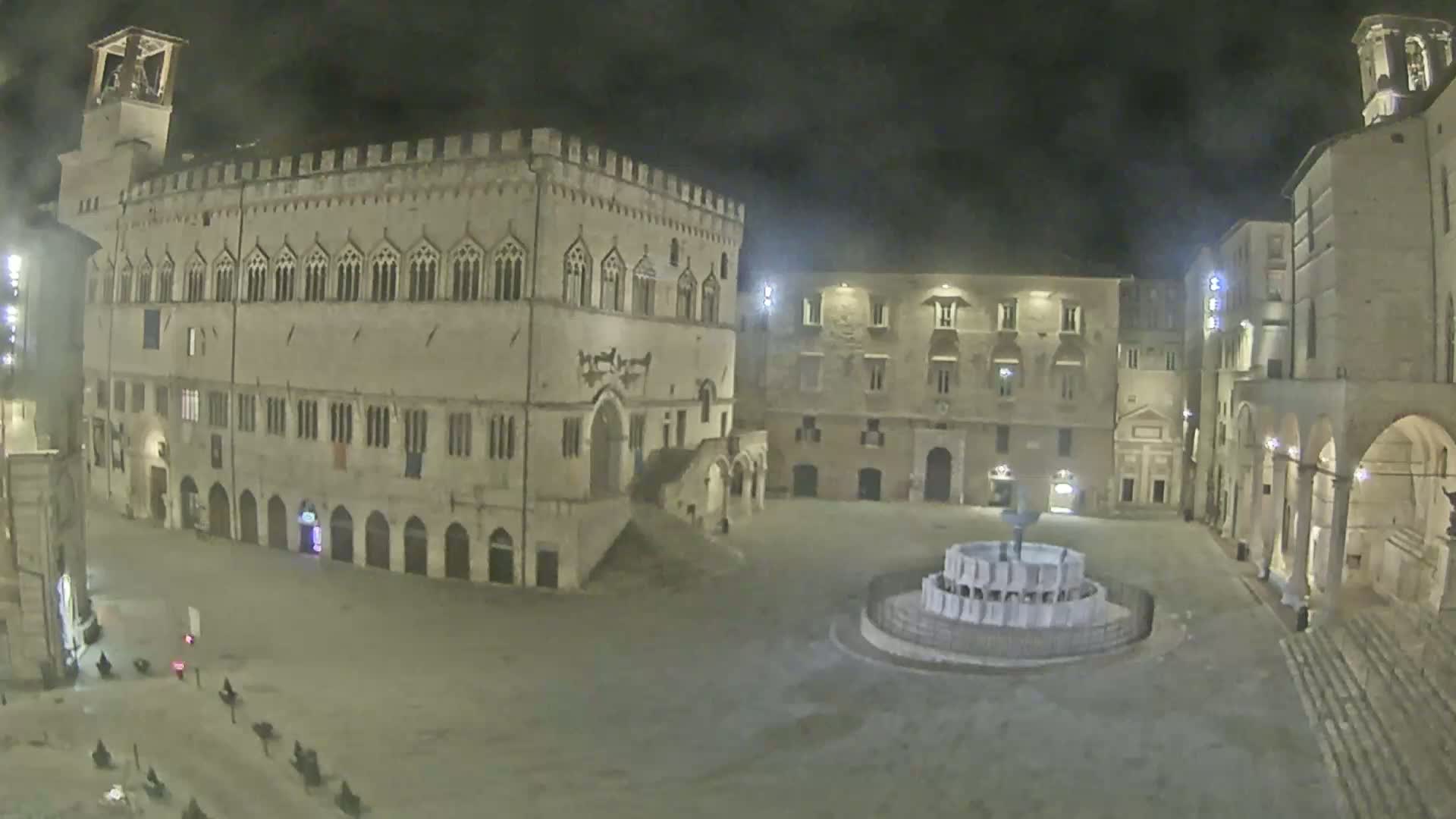 A nighttime view of a large, empty town square with a fountain at its center, surrounded by stone buildings.