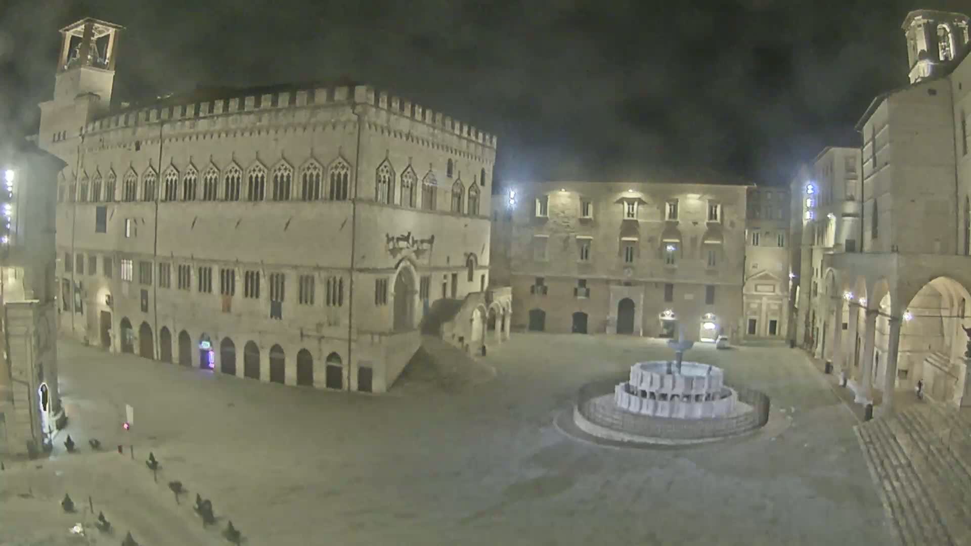 A nighttime view of a large, mostly empty town square with a fountain at its center, surrounded by historic-looking buildings.