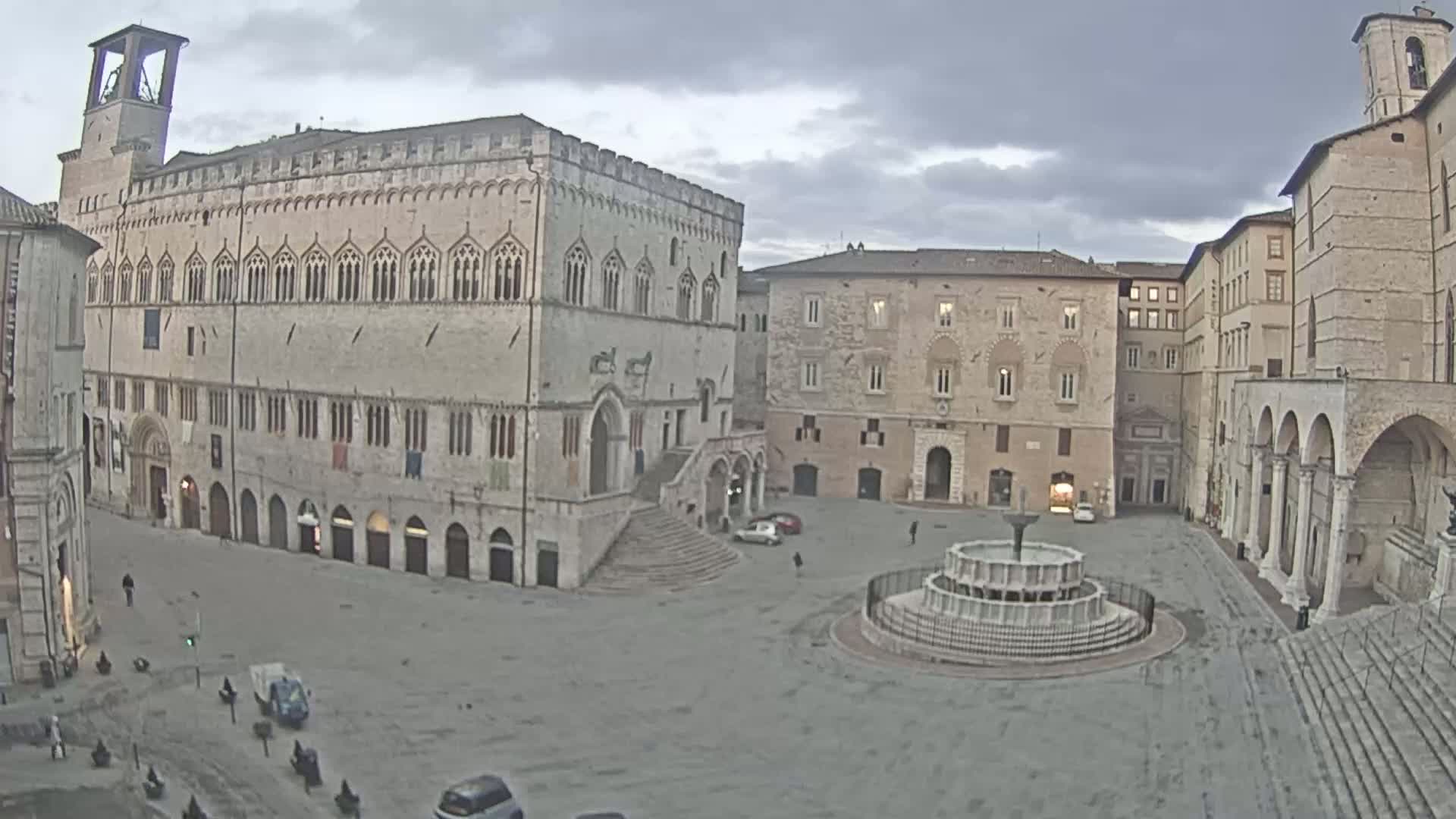 A large, stone plaza with a fountain is surrounded by several historic-looking buildings under an overcast sky.