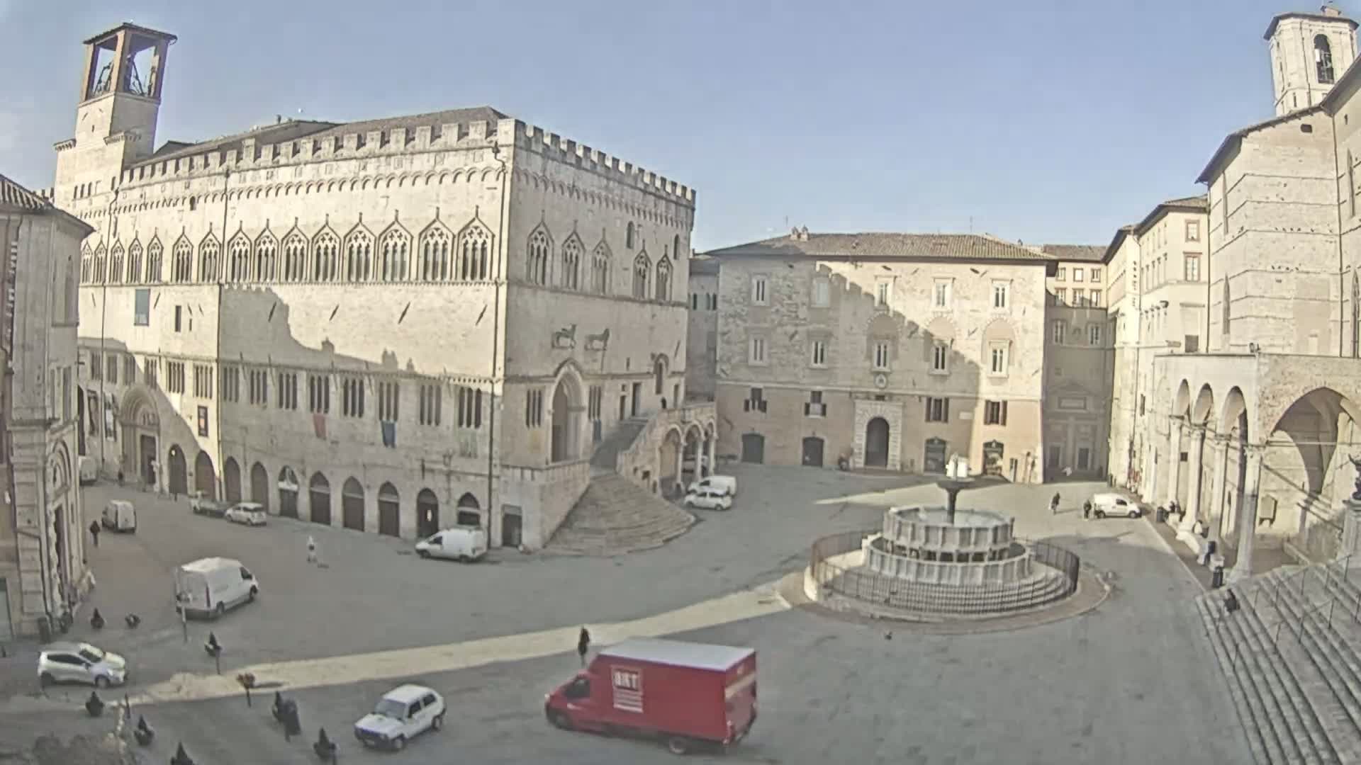 A large, sunlit town square contains a fountain, several vehicles, and historical stone buildings under a clear sky.
