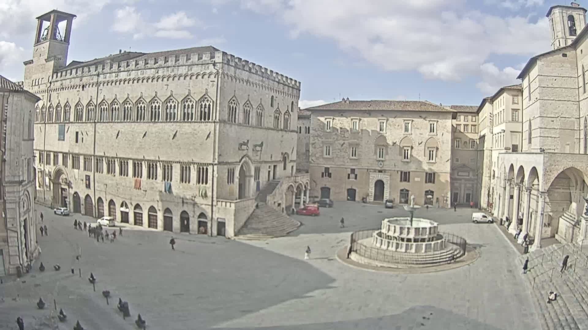 A large, mostly empty town square is centered around a fountain, surrounded by several stone buildings under a partly cloudy sky.