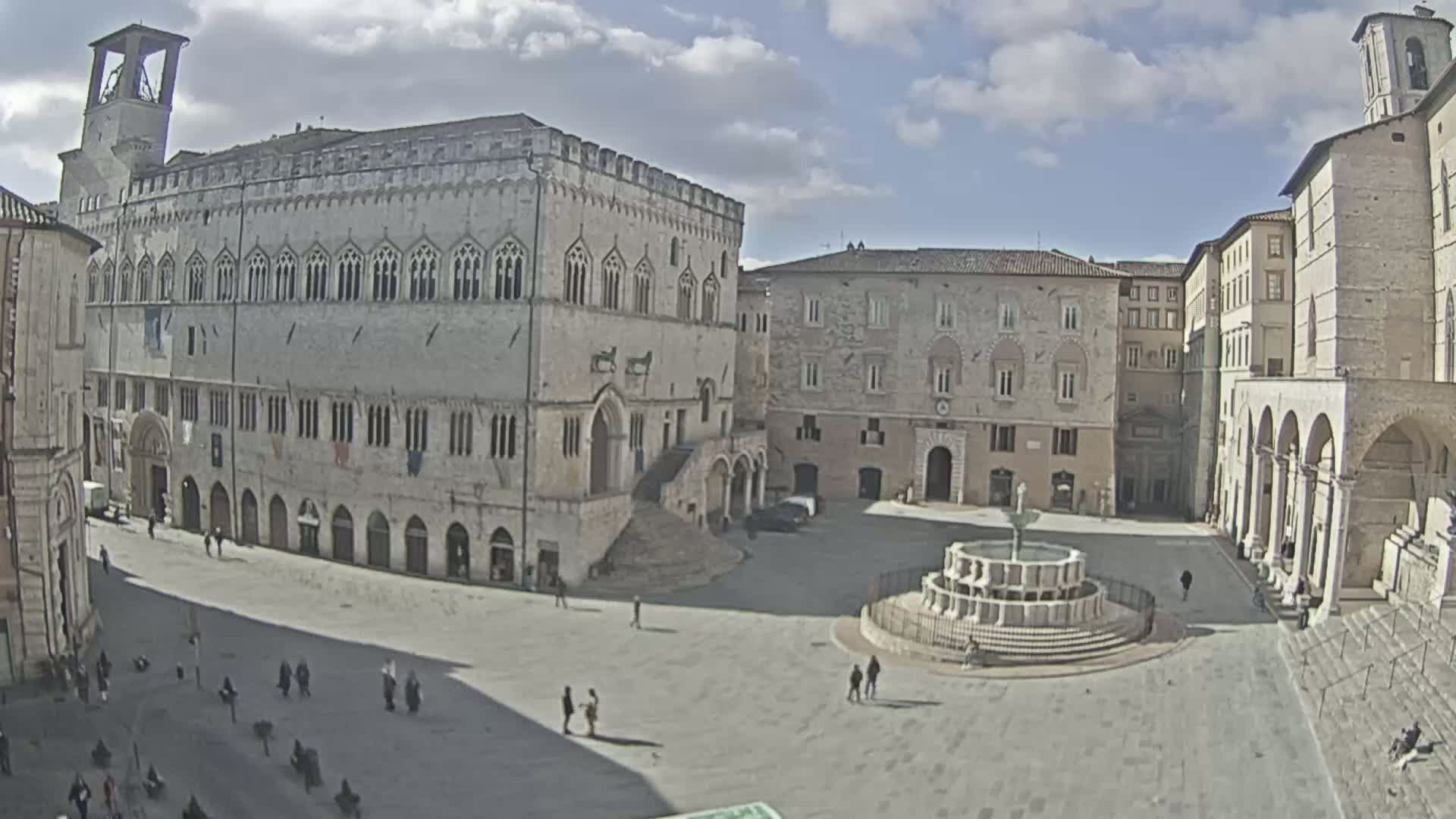 A large, mostly empty town square with several stone buildings, a fountain, and a few people under a partly cloudy sky.