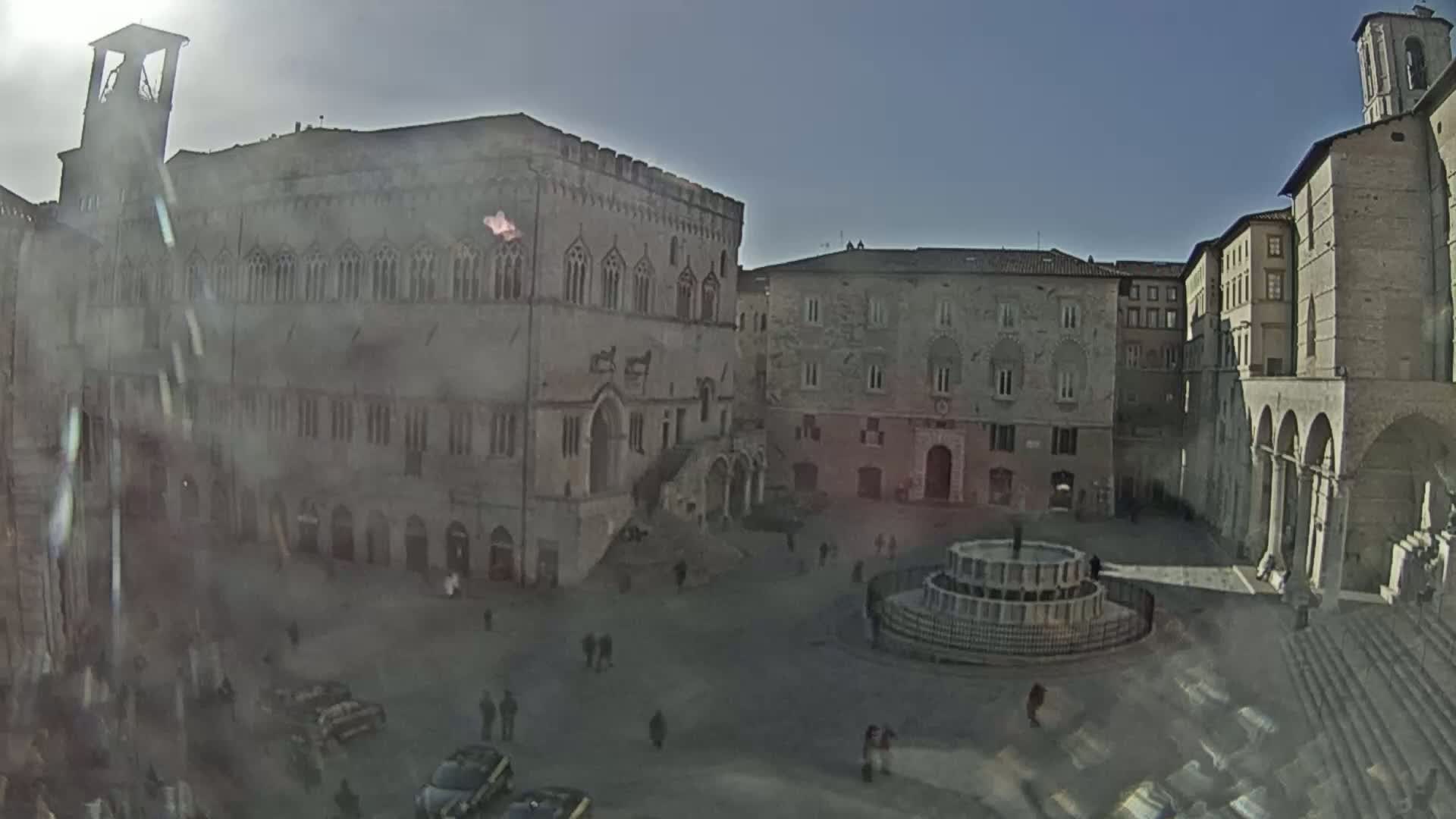 A sunny, hazy day reveals a large town square with several historical buildings, including a palace and a church, and a fountain in the center.