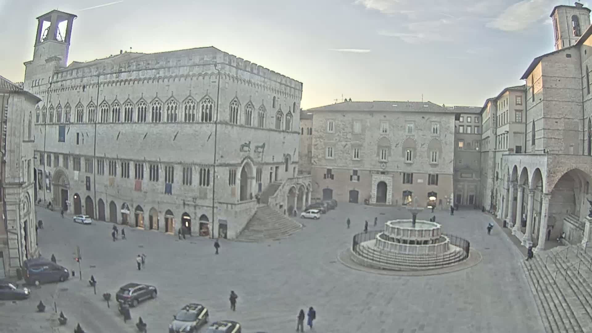 A large, stone plaza with a fountain in the center is surrounded by several historic buildings under a cloudy sky.