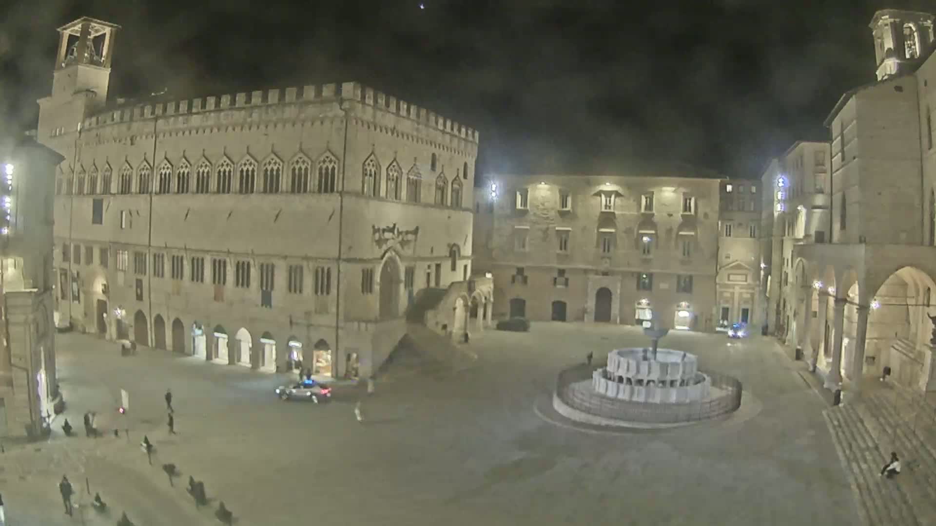 A nighttime view of a large, mostly empty town square, featuring a central fountain, several stone buildings, and a few pedestrians.