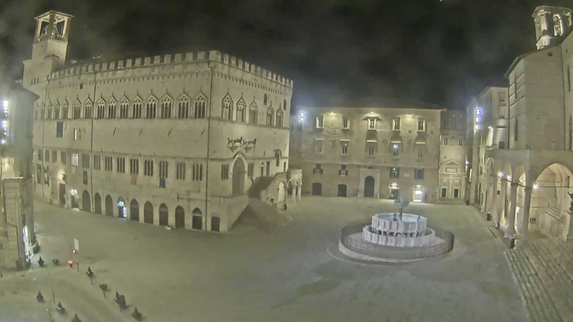A large, mostly empty, stone plaza at night is illuminated, featuring a central fountain and several historic buildings, including a large palace.
