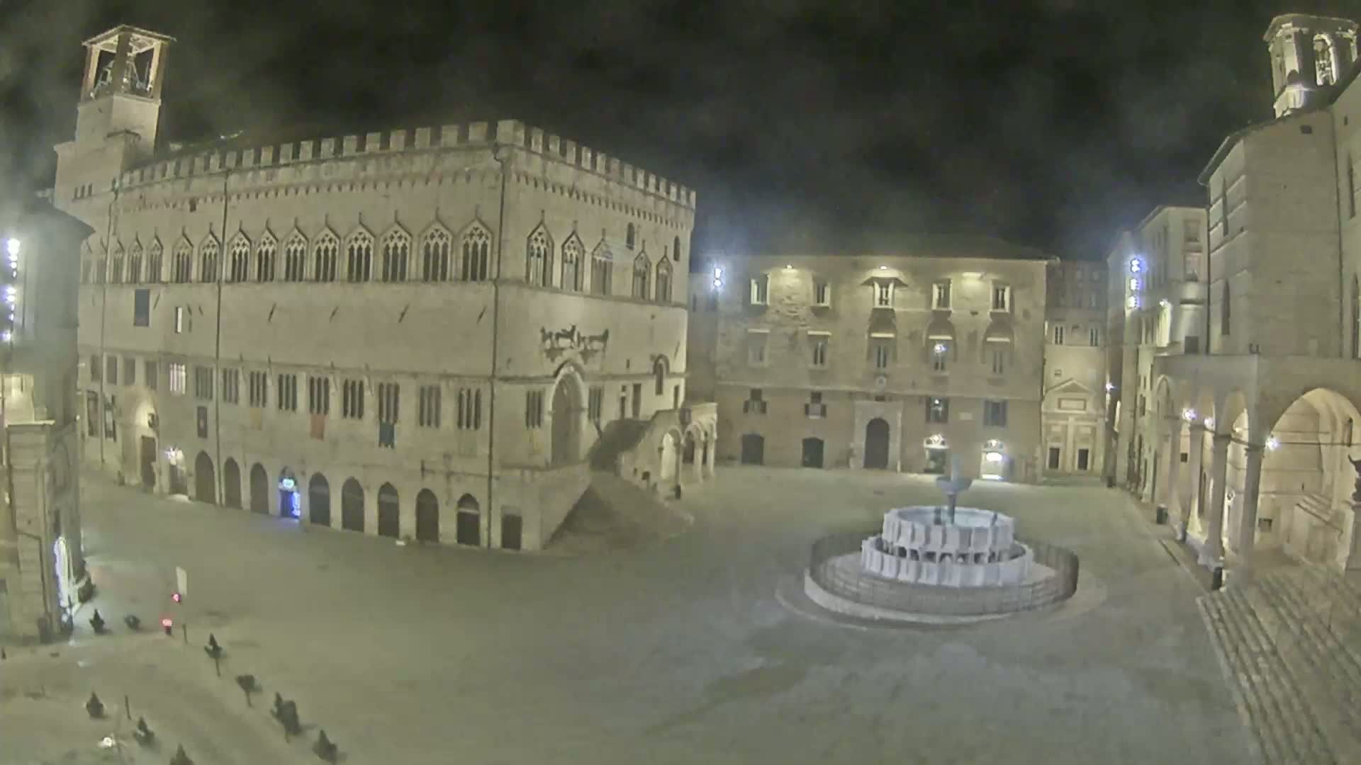 A nighttime, high-angle view of a large, mostly empty town square with several stone buildings, including a large palace, surrounding a central fountain.
