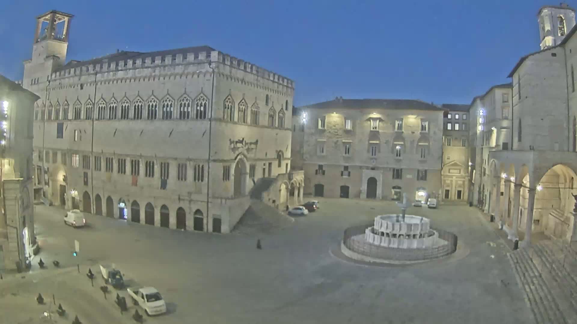 A large, empty town square at twilight is surrounded by historic stone buildings, one of which is a large palace with many windows, and features a circular fountain in the center.