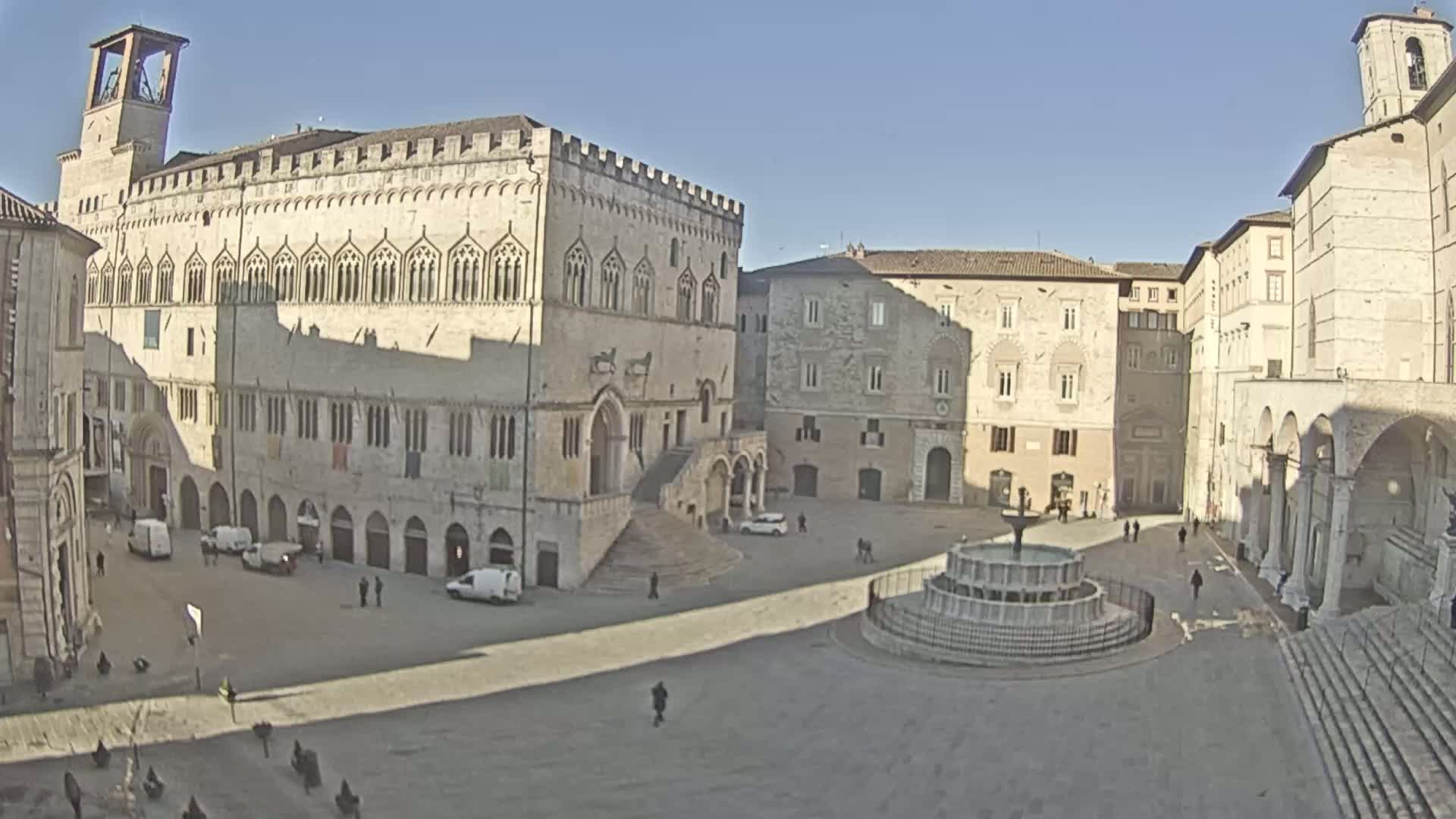 A large, mostly empty town square is surrounded by aged stone buildings, a central fountain, and a few pedestrians under a clear, sunny sky.