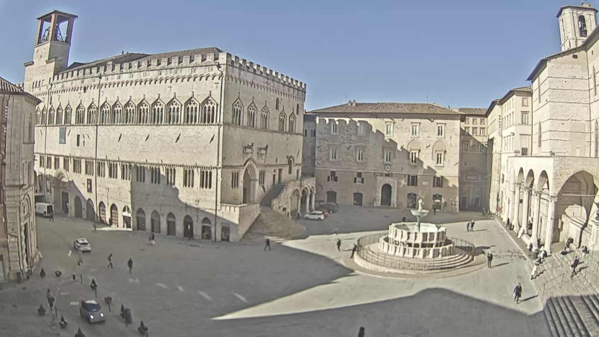 A large town square with a fountain in the center is surrounded by several light-stone buildings under a clear, sunny sky.