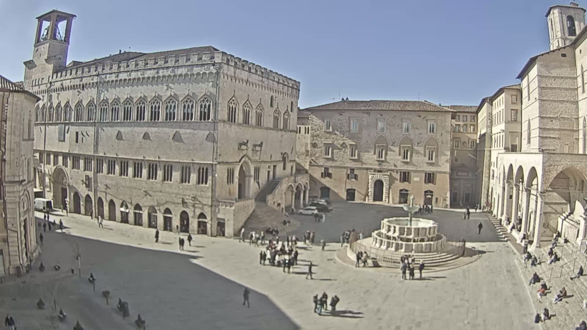 A large, sunny town square is surrounded by medieval-style buildings, featuring a central fountain and a scattering of people.