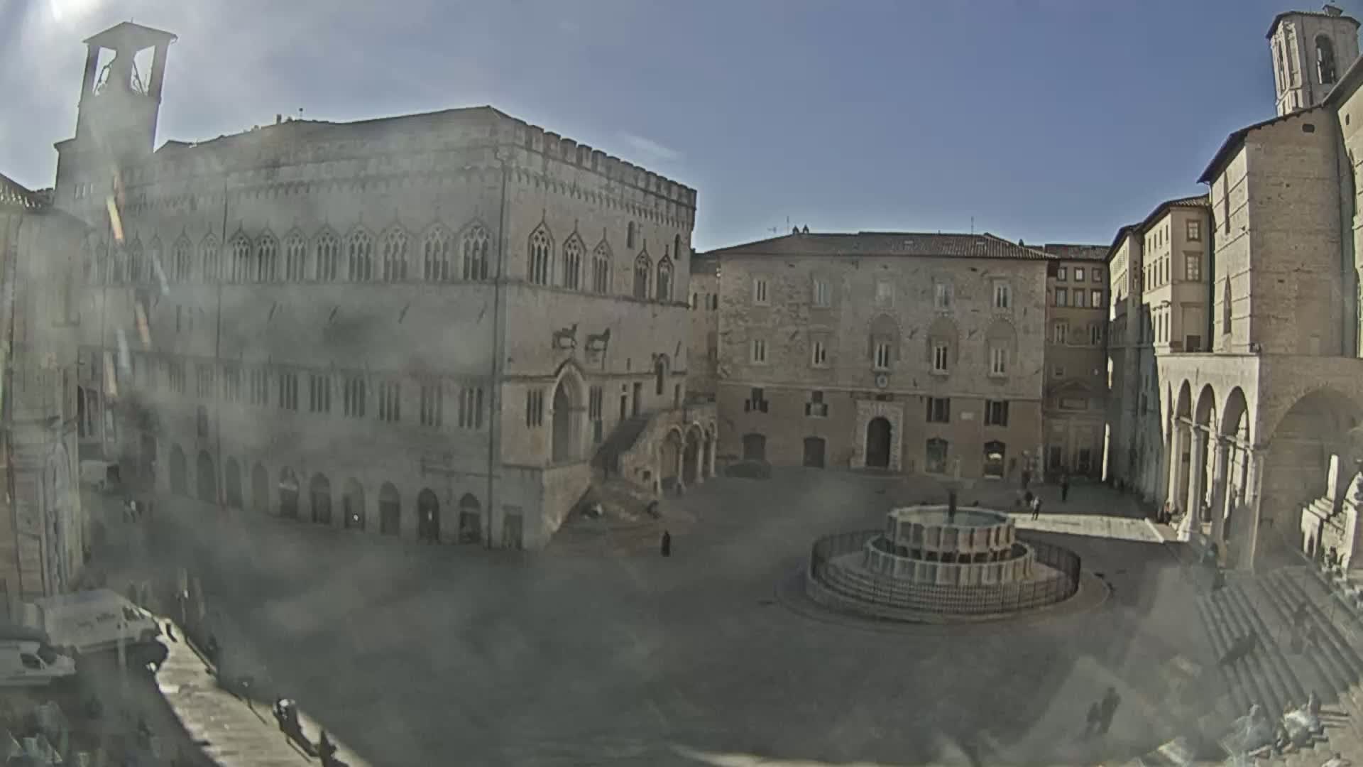 A sunny day reveals a large town square surrounded by historical buildings, including a prominent palace and a church, with a central fountain.