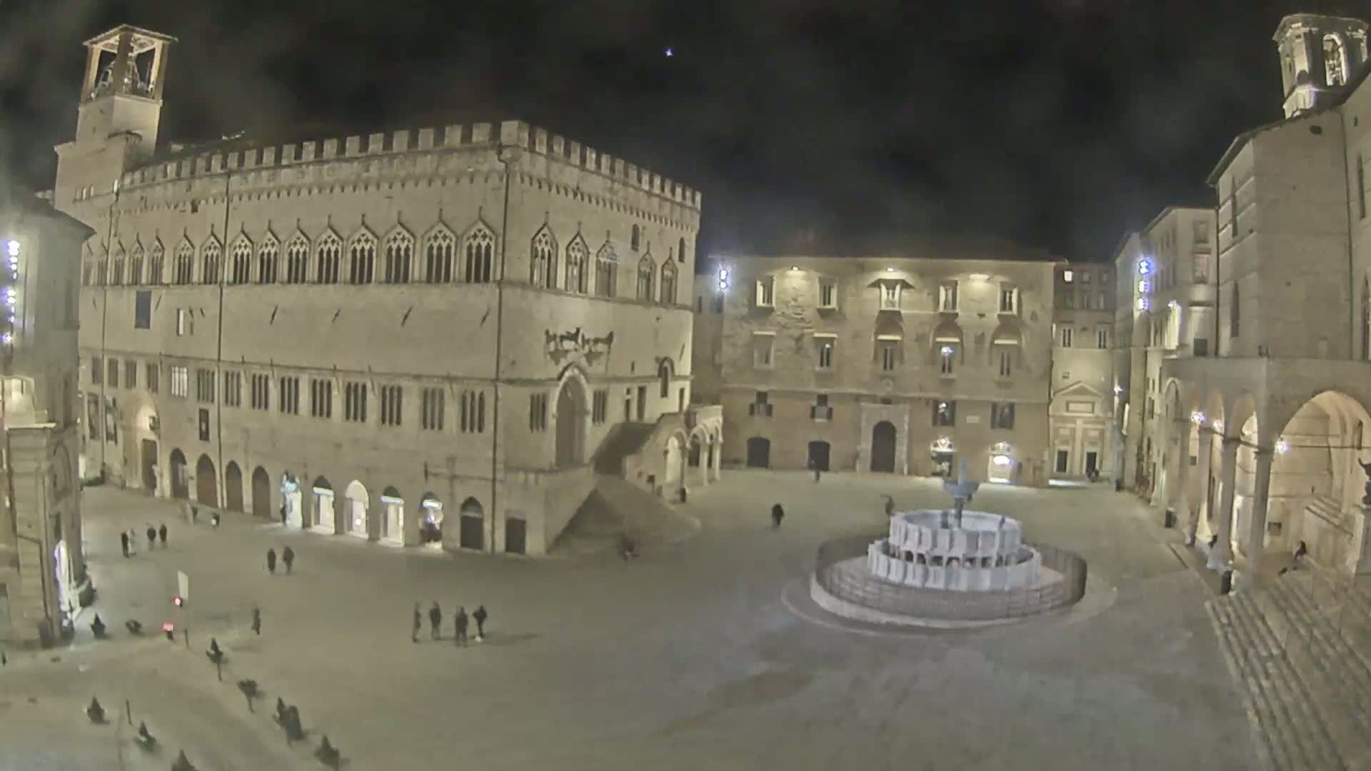 A large, empty town square at night is surrounded by medieval-style buildings, with a fountain at its center under a clear, dark sky.