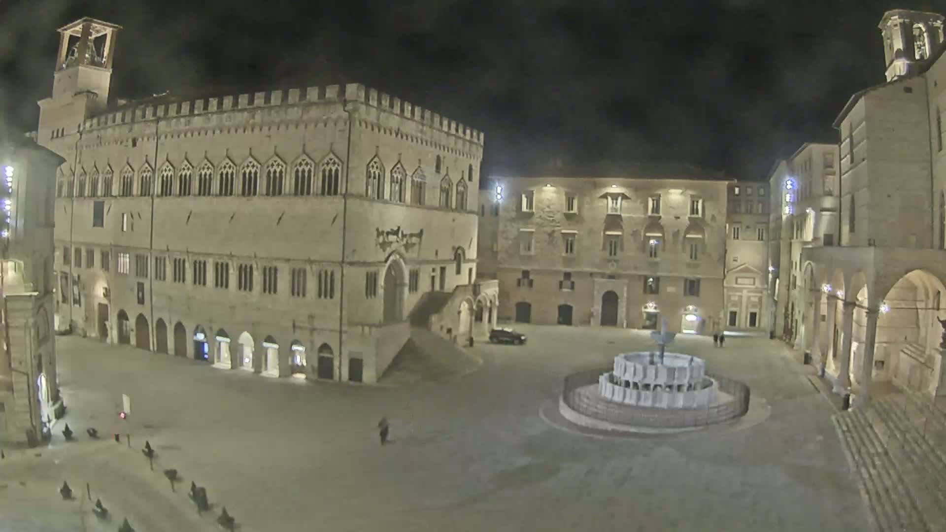 A large, mostly empty town square at night, featuring a fountain and several stone buildings, one of which is a large palace.