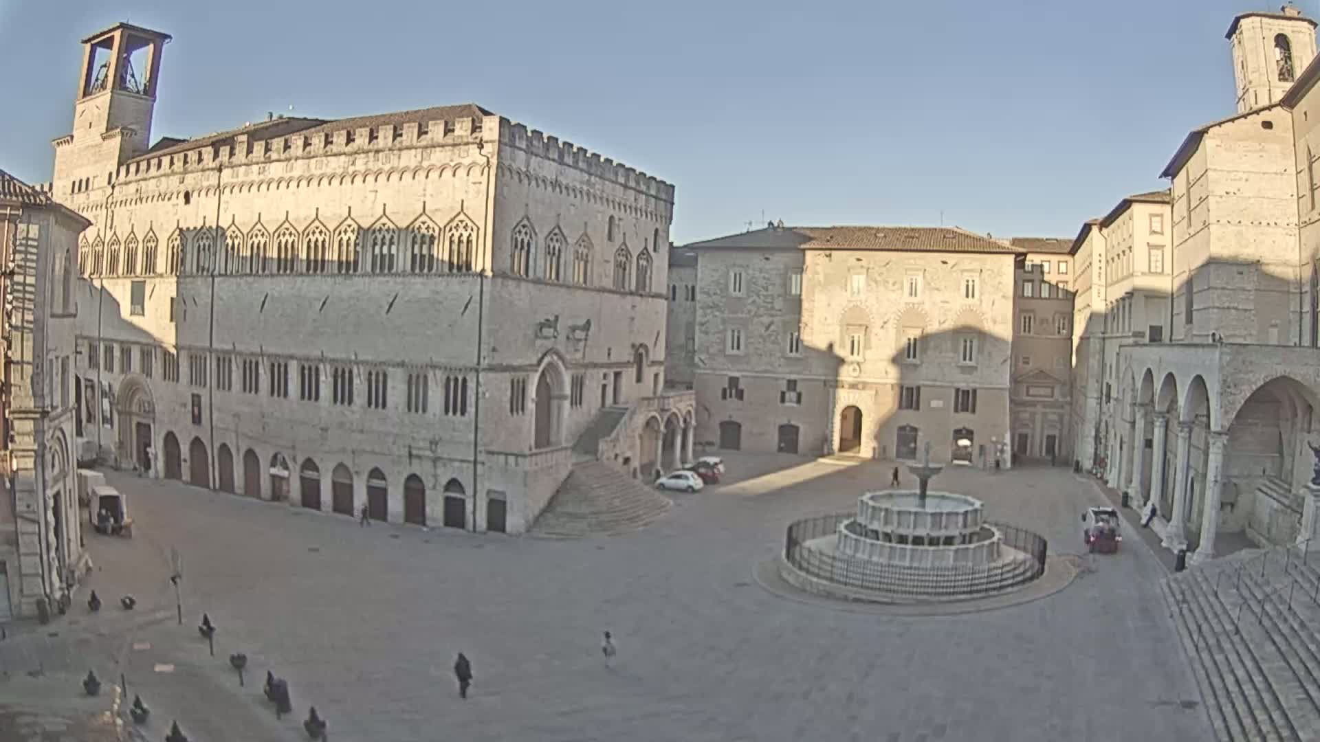 A large, mostly empty town square with a fountain at its center, surrounded by ancient stone buildings under a clear, sunny sky.