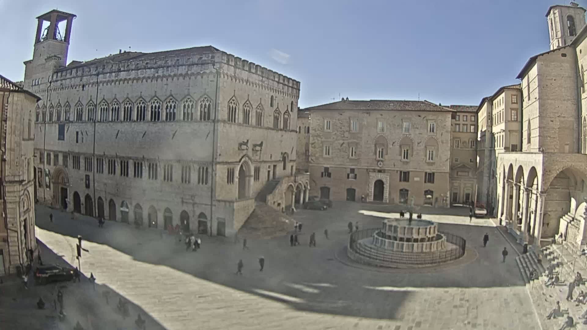 A sunny day reveals a large town square with several stone buildings, including a prominent palace, and a fountain in the center.