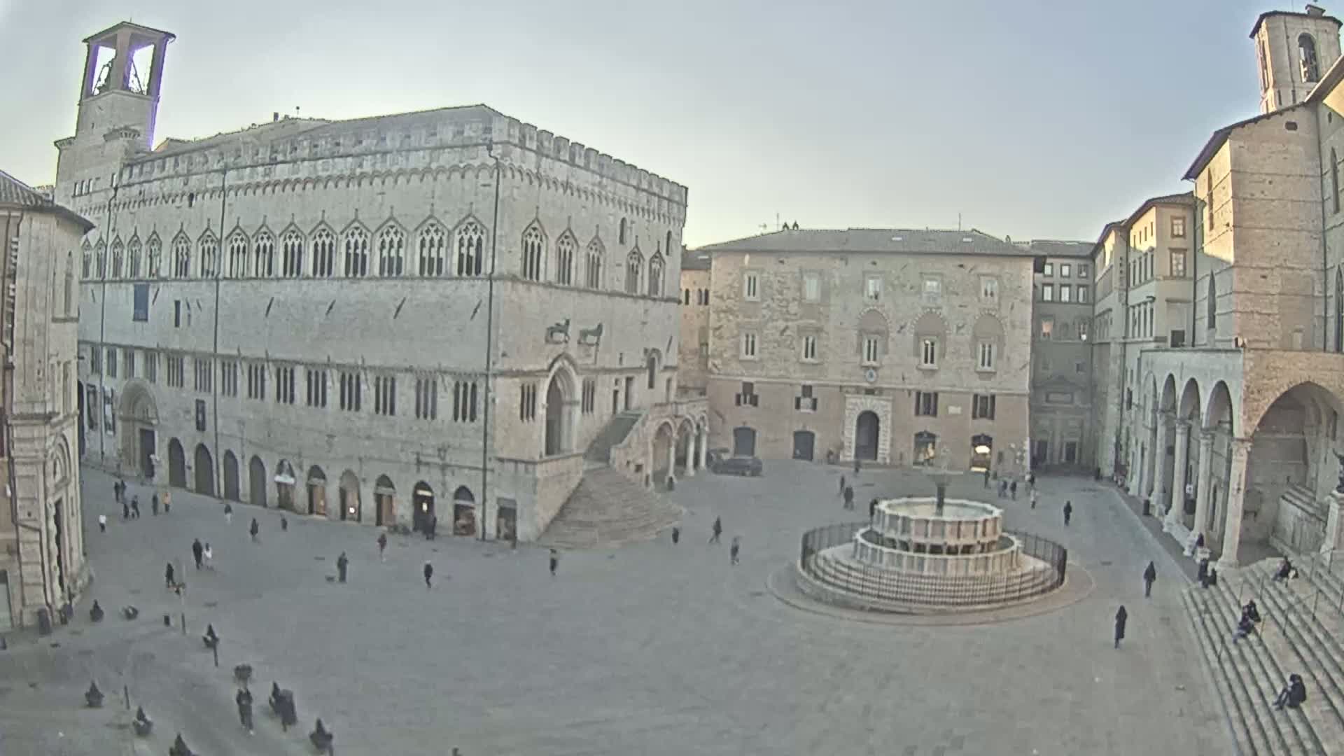 A large town square with several stone buildings, a central fountain, and a few scattered people under a bright, clear sky.