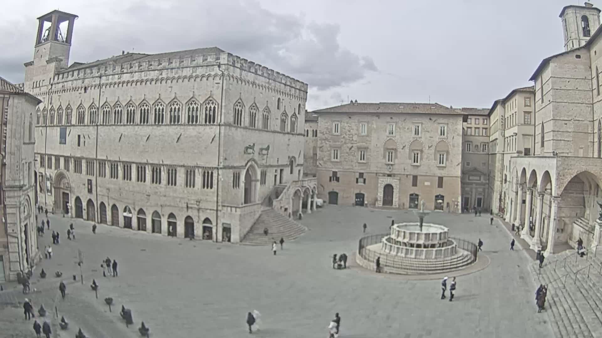 A large, mostly empty town square is surrounded by aged stone buildings under an overcast sky, featuring a central fountain.