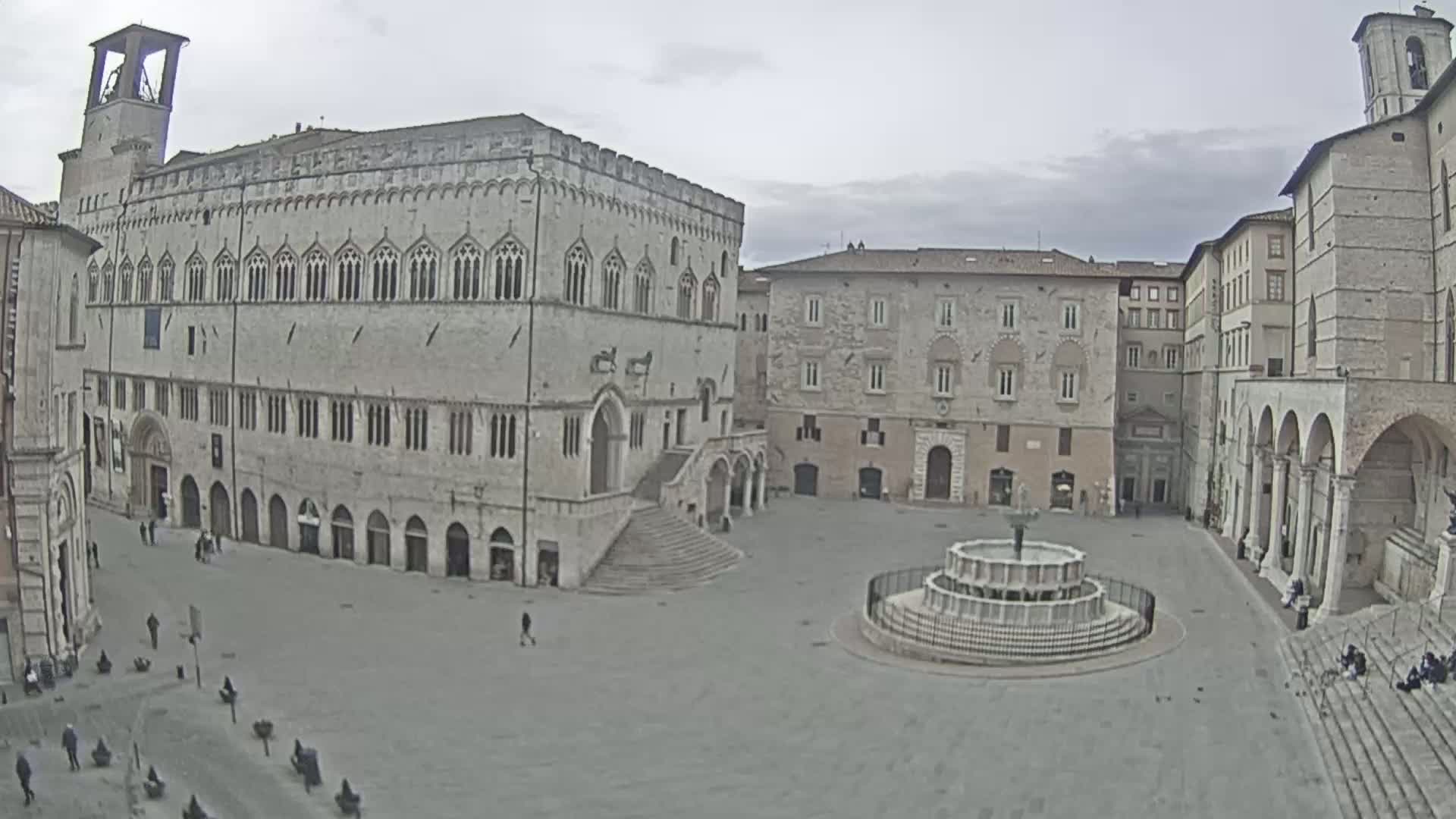 A large, empty town square with a fountain in the center is surrounded by several historic stone buildings under an overcast sky.